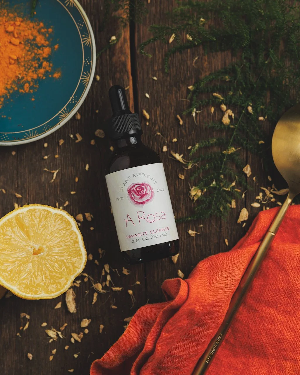 A dark amber bottle labeled 'A Rosa' with a pink flower illustration, a lemon half, a blue dish with golden powder, green foliage, and a gold spoon with an orange cloth on a wooden surface.