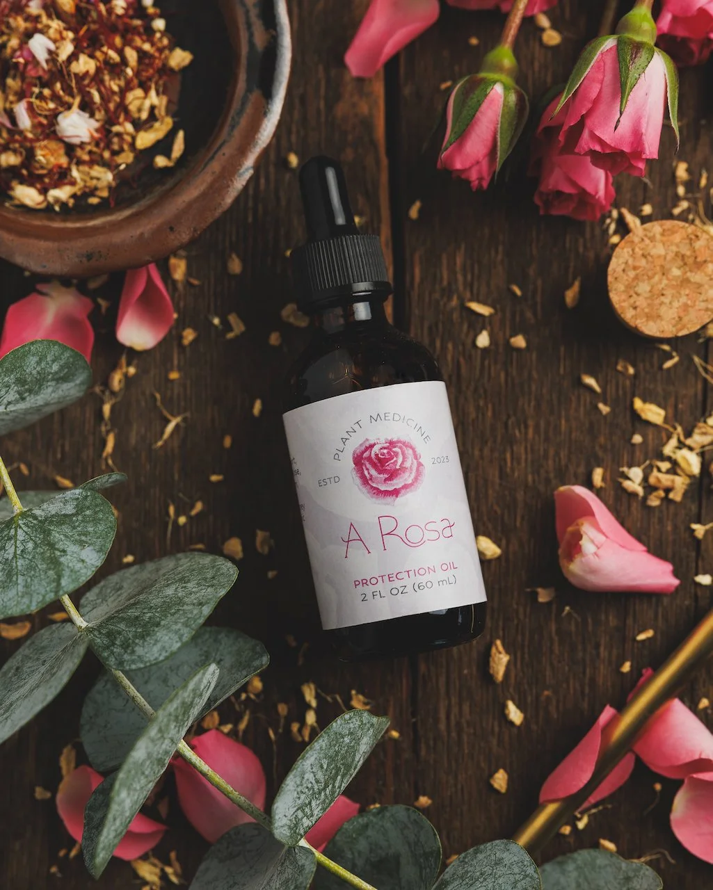 A small dark bottle labeled 'A Rosa Protection Oil,' surrounded by rose petals, pink roses, eucalyptus leaves, a bowl of dried rose petals, and a small round wooden object on a wooden surface.