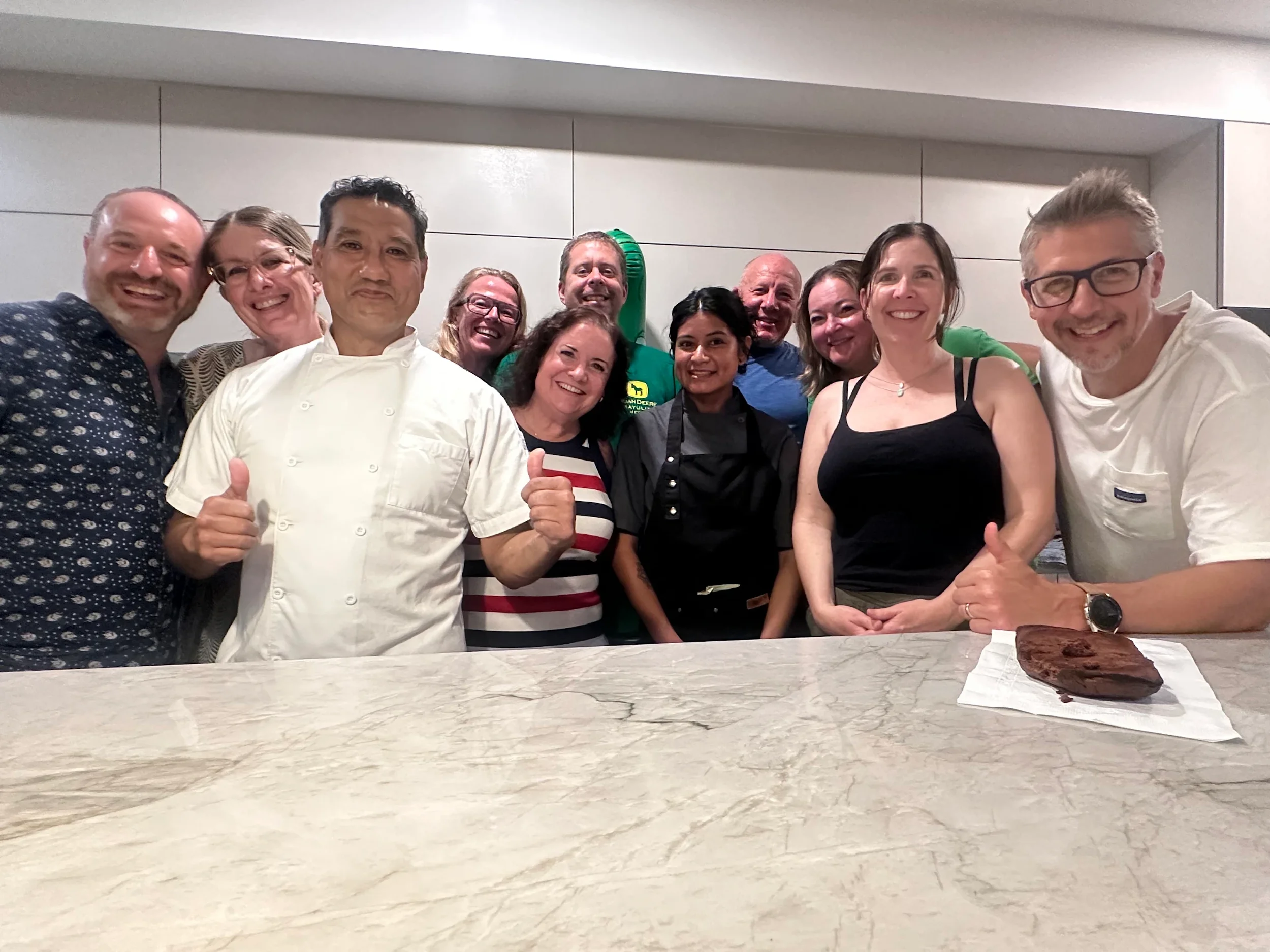 A group of twelve people, including a chef in a white uniform and people with aprons, smiling and posing together in a kitchen or restaurant setting. There is a marble countertop with a cake and a piece of cake in the foreground.