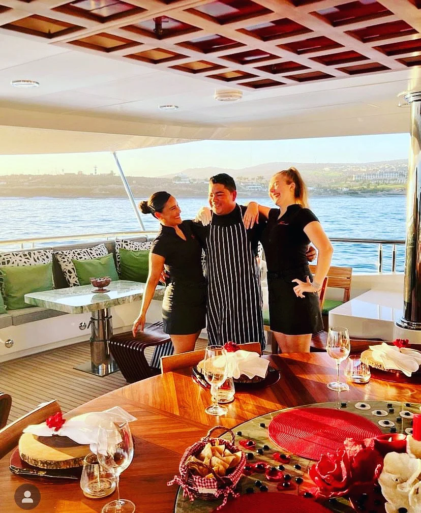 Three people standing on a boat deck, a male chef in the center with two women on either side, smiling and posing with arms around each other, overlooking water with distant shoreline.
