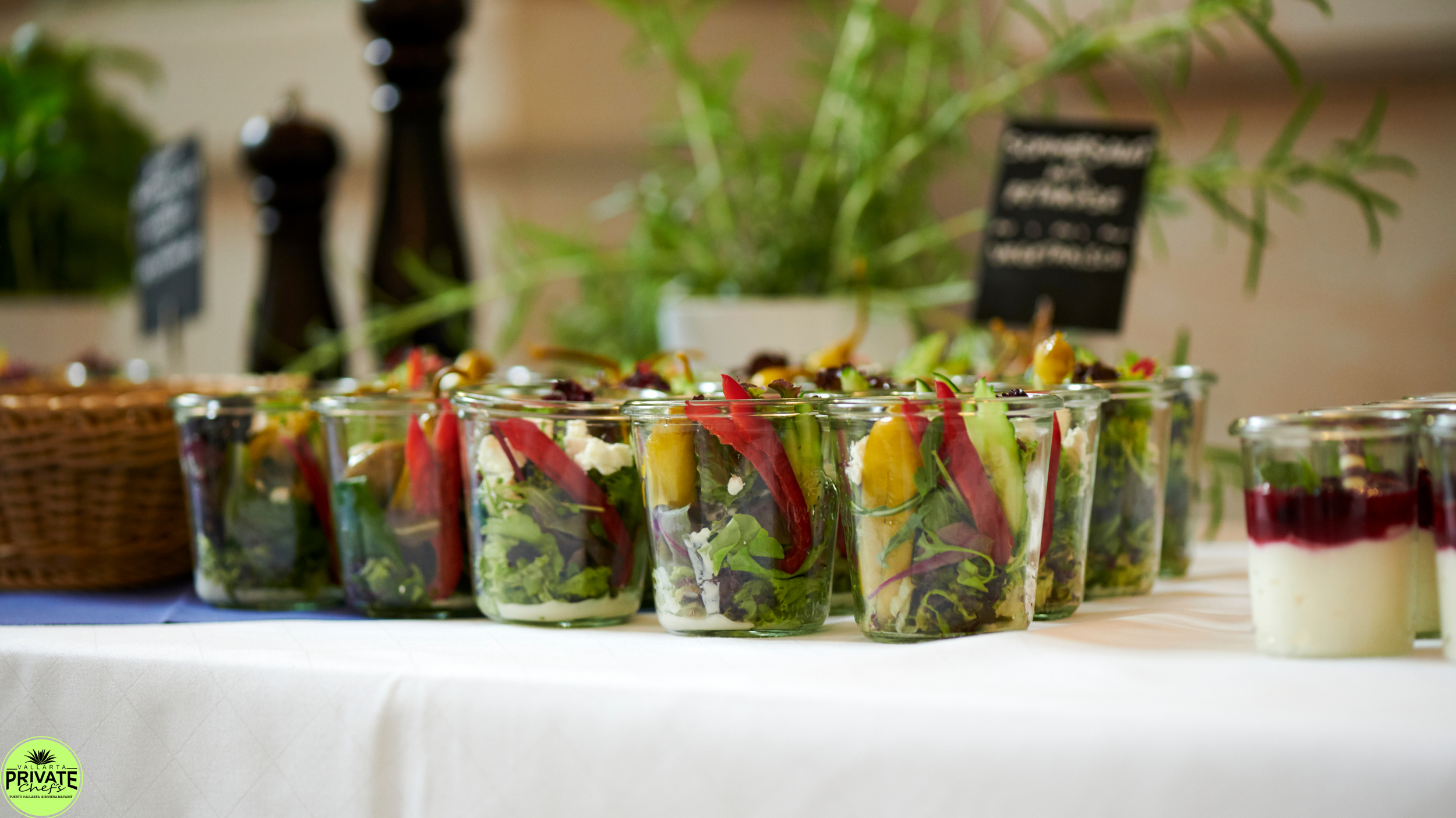 Rows of salad in clear glass cups on a table with a white tablecloth, with other food items and blurred background.