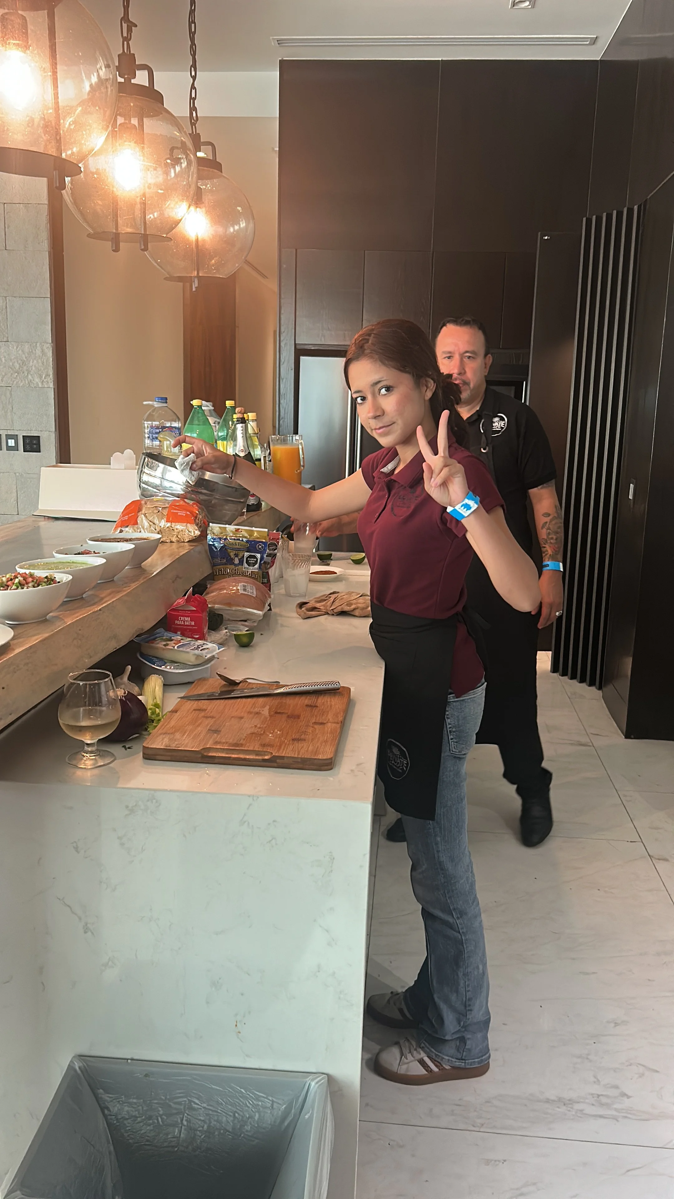 A woman in a maroon shirt and apron making a peace sign with her right hand, standing at a kitchen counter with food ingredients, while a man behind her looks on. The kitchen has modern lighting and dark cabinets.