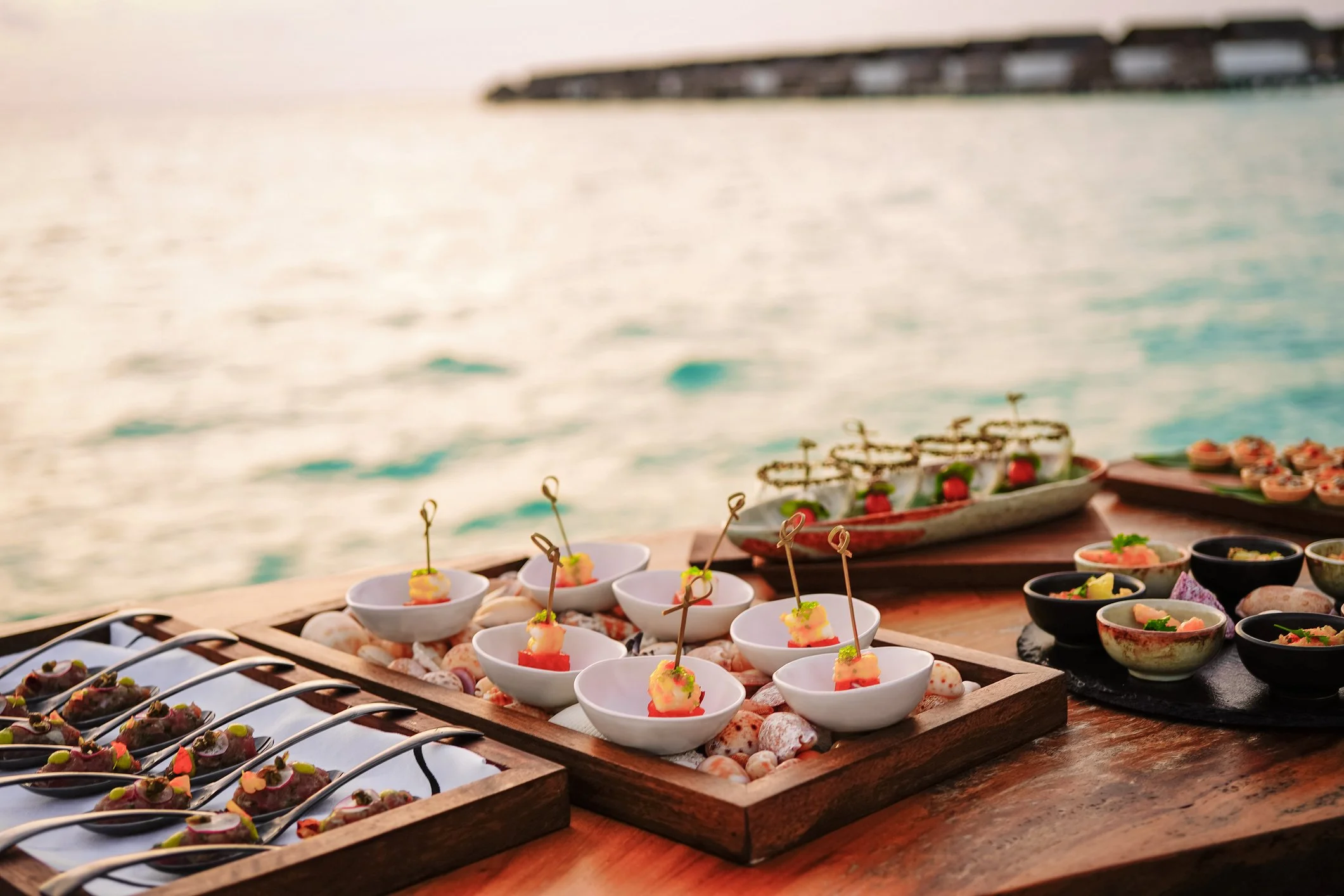 Assorted appetizers on a wooden table by the water, with the sea and a distant shoreline in the background.