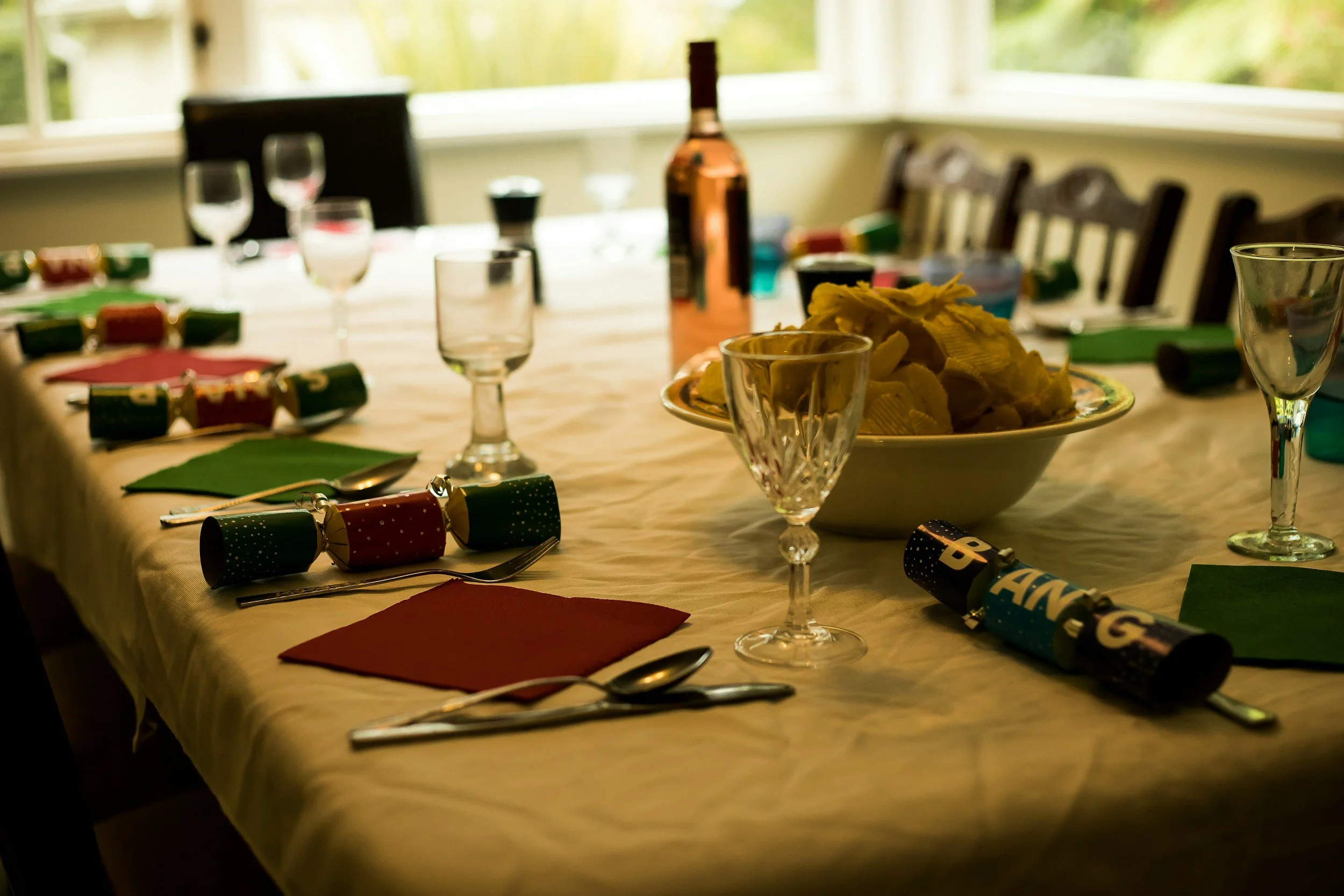 A decorated dining table set for a celebration with party hats, napkins, glasses, a bowl of chips, and a bottle, with chairs and a window in the background.