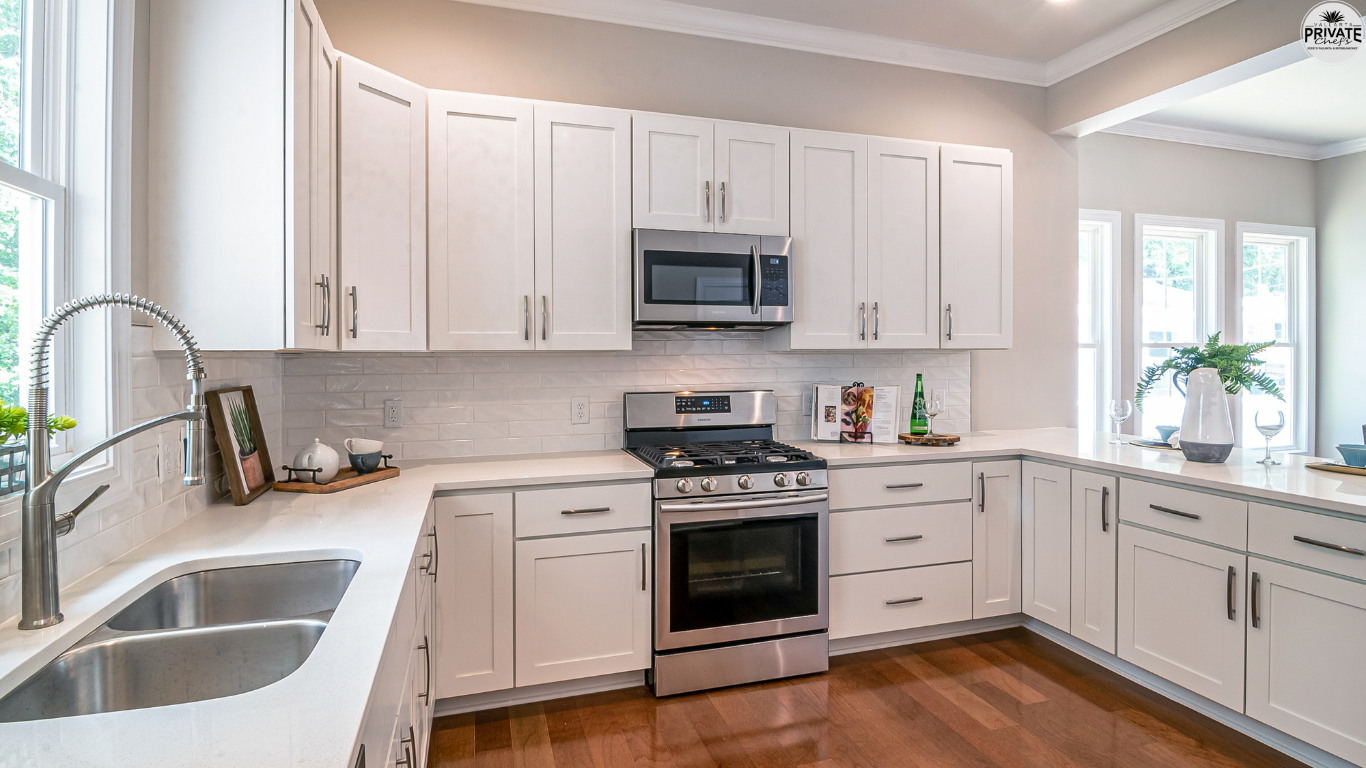 Modern white kitchen with stainless steel appliances, including a microwave, oven, and stove, set against a tiled backsplash. The kitchen features white cabinetry, a window above the sink, and wooden floor.