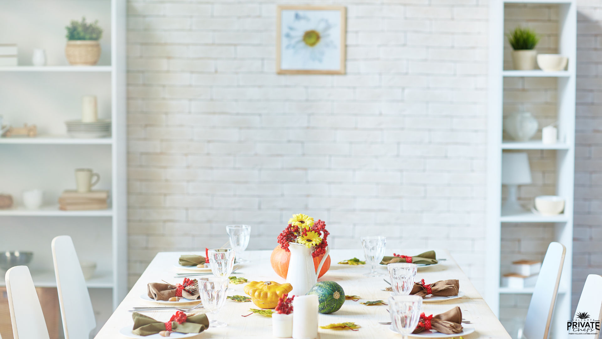 Dining table decorated with autumn-themed centerpiece including a white pitcher with yellow and red flowers, pumpkins, and candles, set in a bright, modern dining room with white shelving and a brick wall.