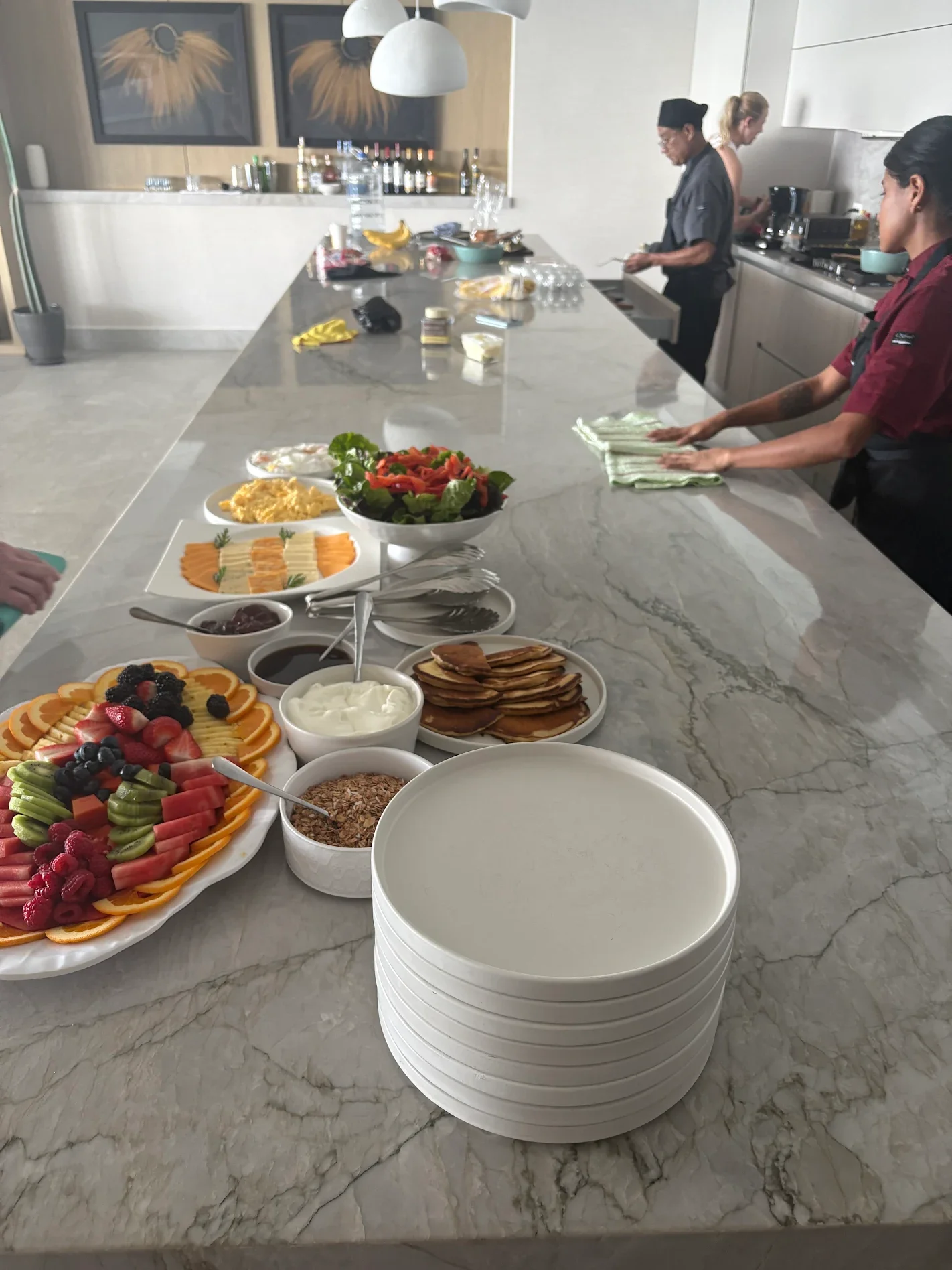 Buffet table with fruit, pancakes, yogurt, syrup, and other breakfast items in a modern kitchen with two staff members preparing food.