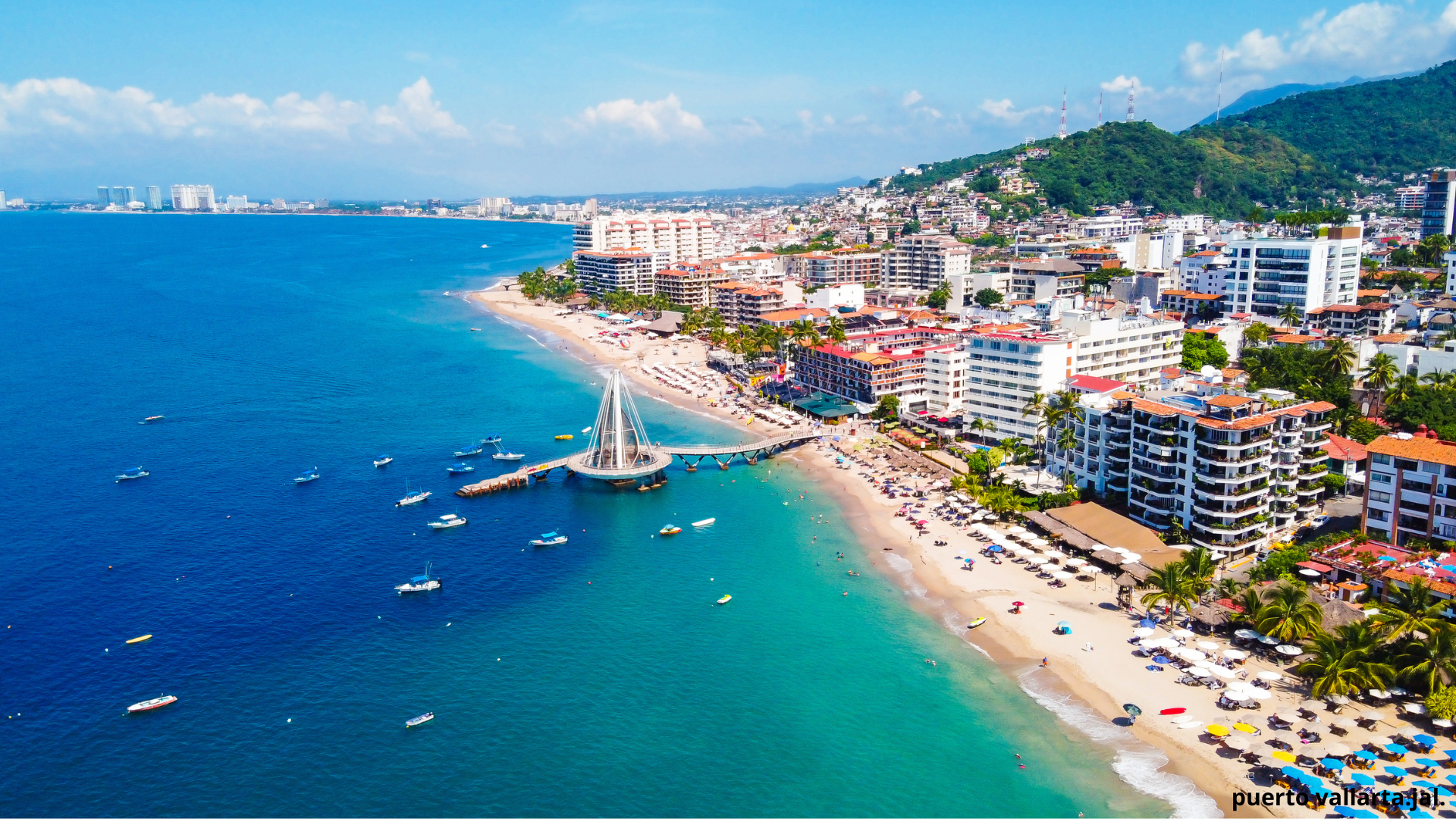 Aerial view of a coastal city with a beach, boats in the water, high-rise buildings, and a lush green hill with communication towers.