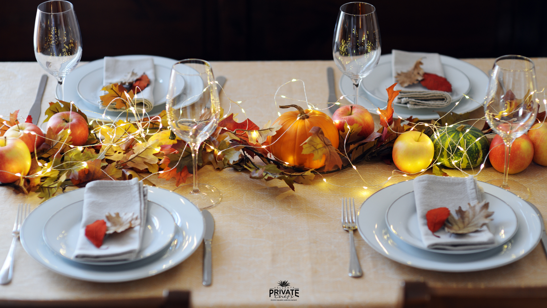 Table decorated for fall or Thanksgiving with apples, pumpkins, autumn leaves, and string lights, set with white plates, napkins, and glasses.