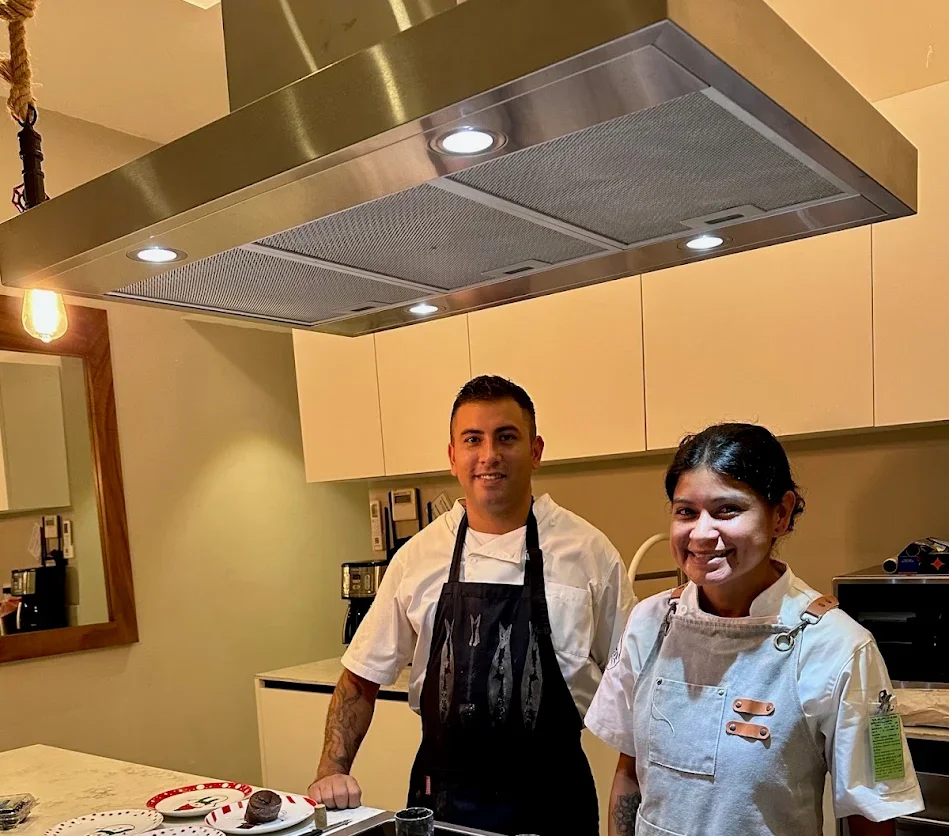 Two chefs in white uniforms standing in a kitchen with a large stainless steel range hood above them, smiling at the camera.