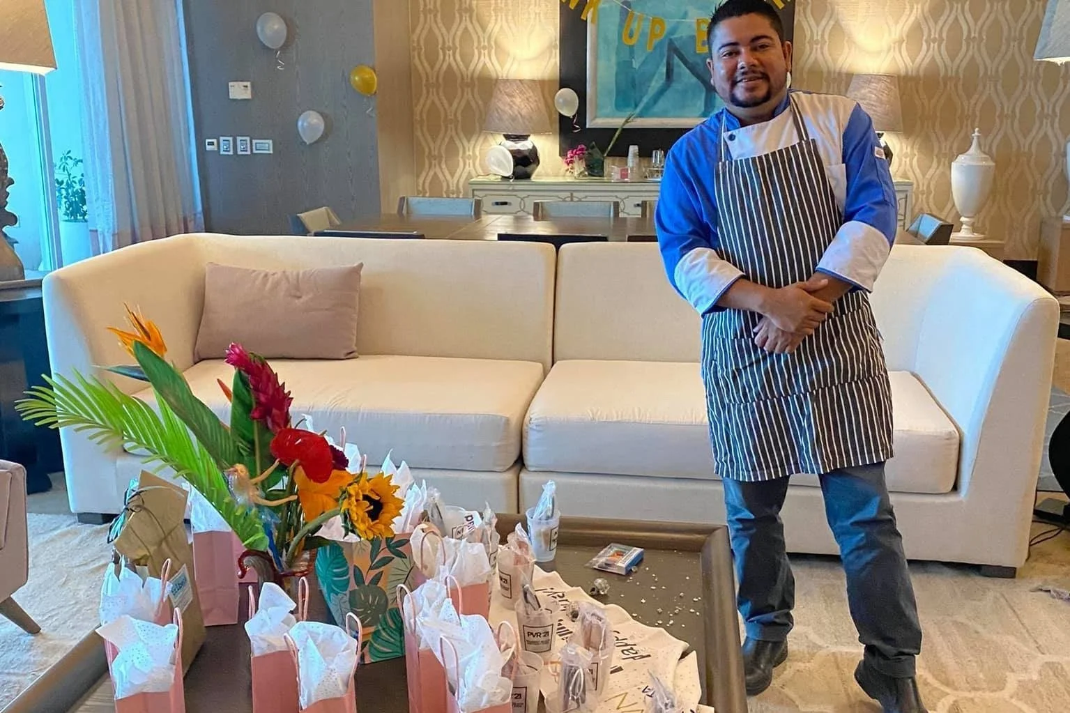 A man in a chef uniform and striped apron standing in a living room with a sofa, a coffee table with flower arrangements and party favor bags, and a decorated wall with balloons in the background.