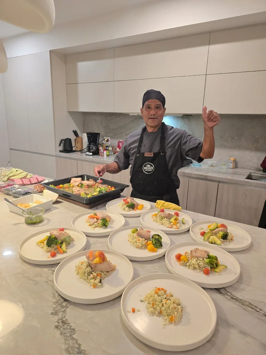 Chef standing in a kitchen giving a thumbs up, with prepared plates of food including rice, vegetables, and chicken on a marble countertop.
