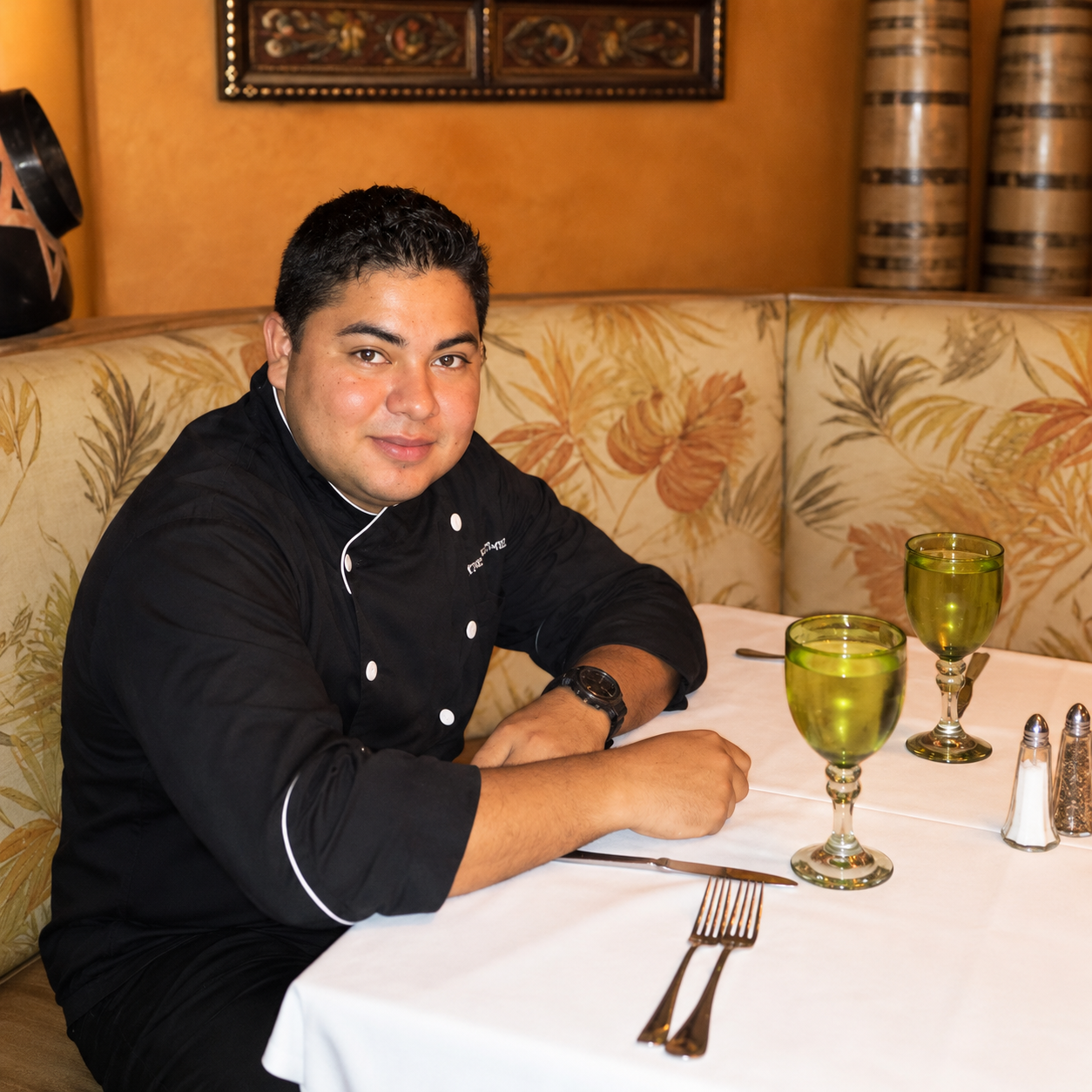 A male chef in a black chef's coat sitting at a table in a restaurant, with two green wine glasses, silverware, and salt and pepper shakers on the table.