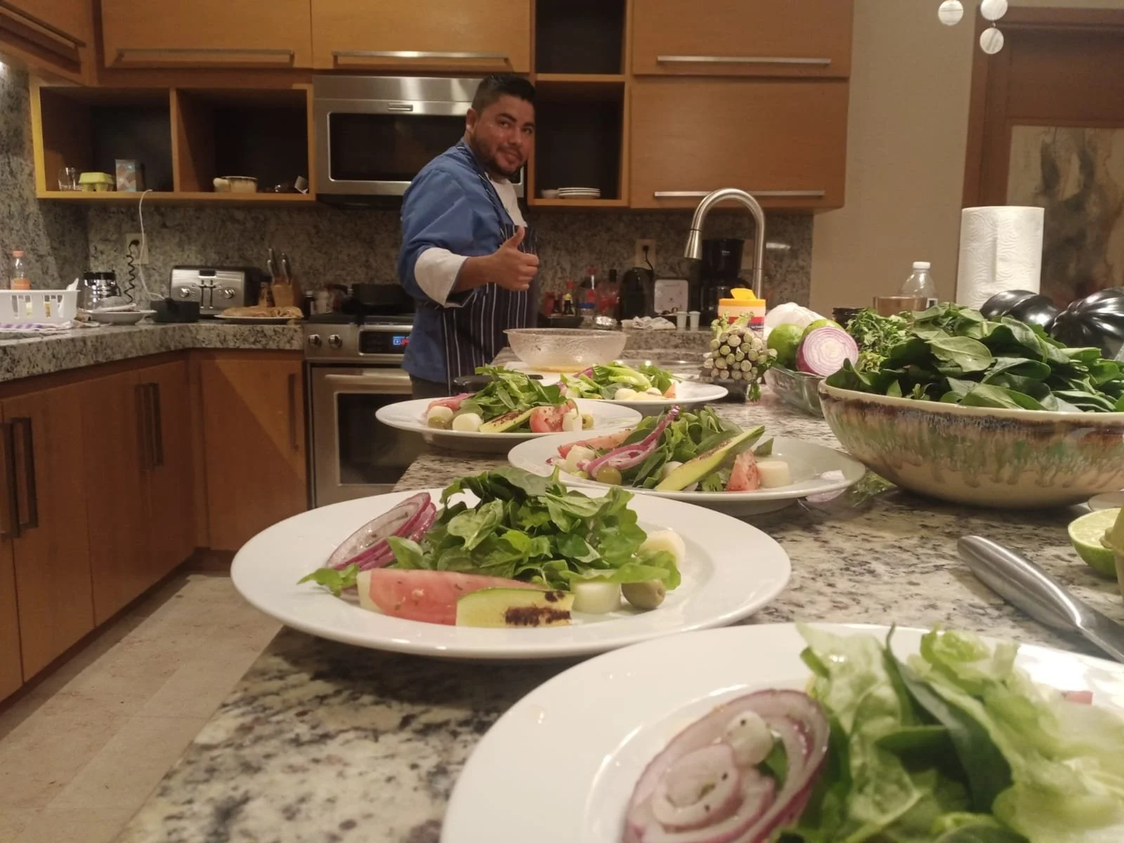 A man in a kitchen giving a thumbs-up next to multiple plates of salad with vegetables like lettuce, tomatoes, onions, and cucumbers on a granite countertop.