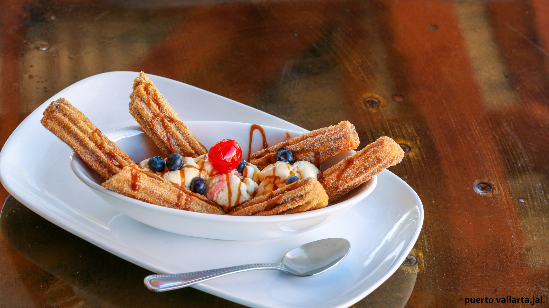A dessert with churros with caramel sauce, ice cream, blueberries, and maraschino cherry in a white bowl on a wooden table.