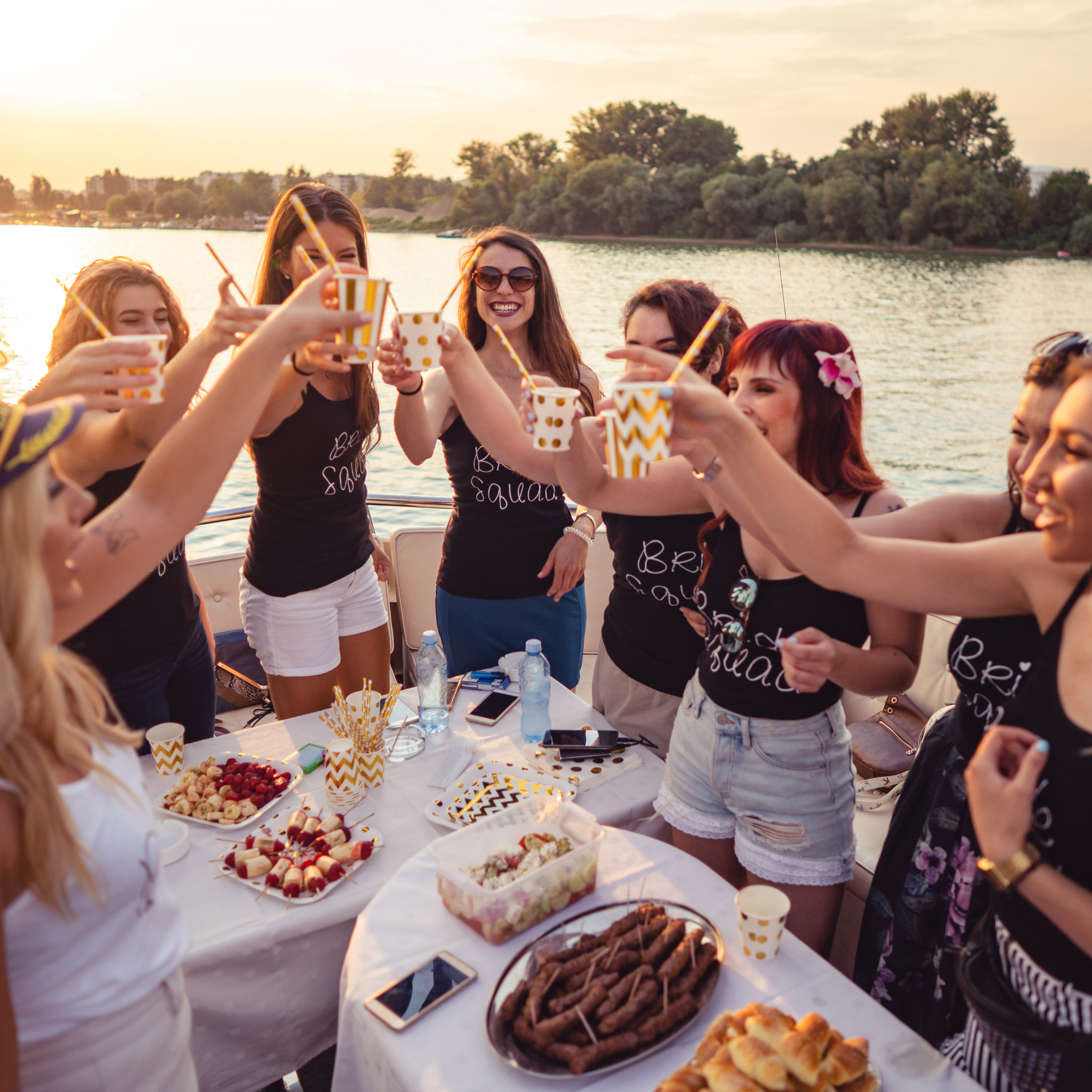 Group of young women enjoying a celebration with food and drinks on a boat by the water during sunset