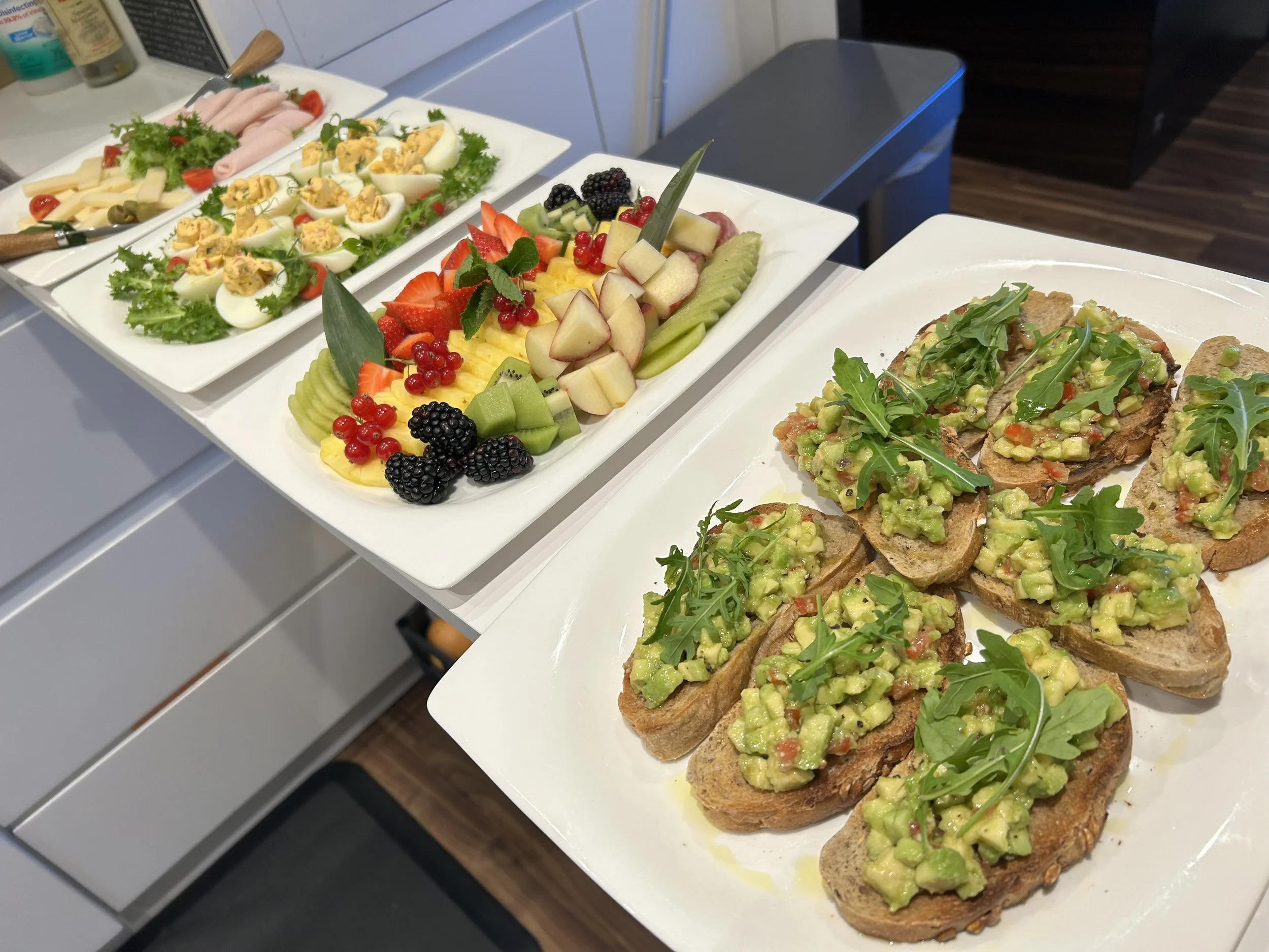 Assorted dishes on white rectangular plates, including deviled eggs with paprika, a fruit platter with strawberries, kiwi, watermelon, honeydew, red currants, blackberries, and apple slices, and slices of bread topped with guacamole and arugula.