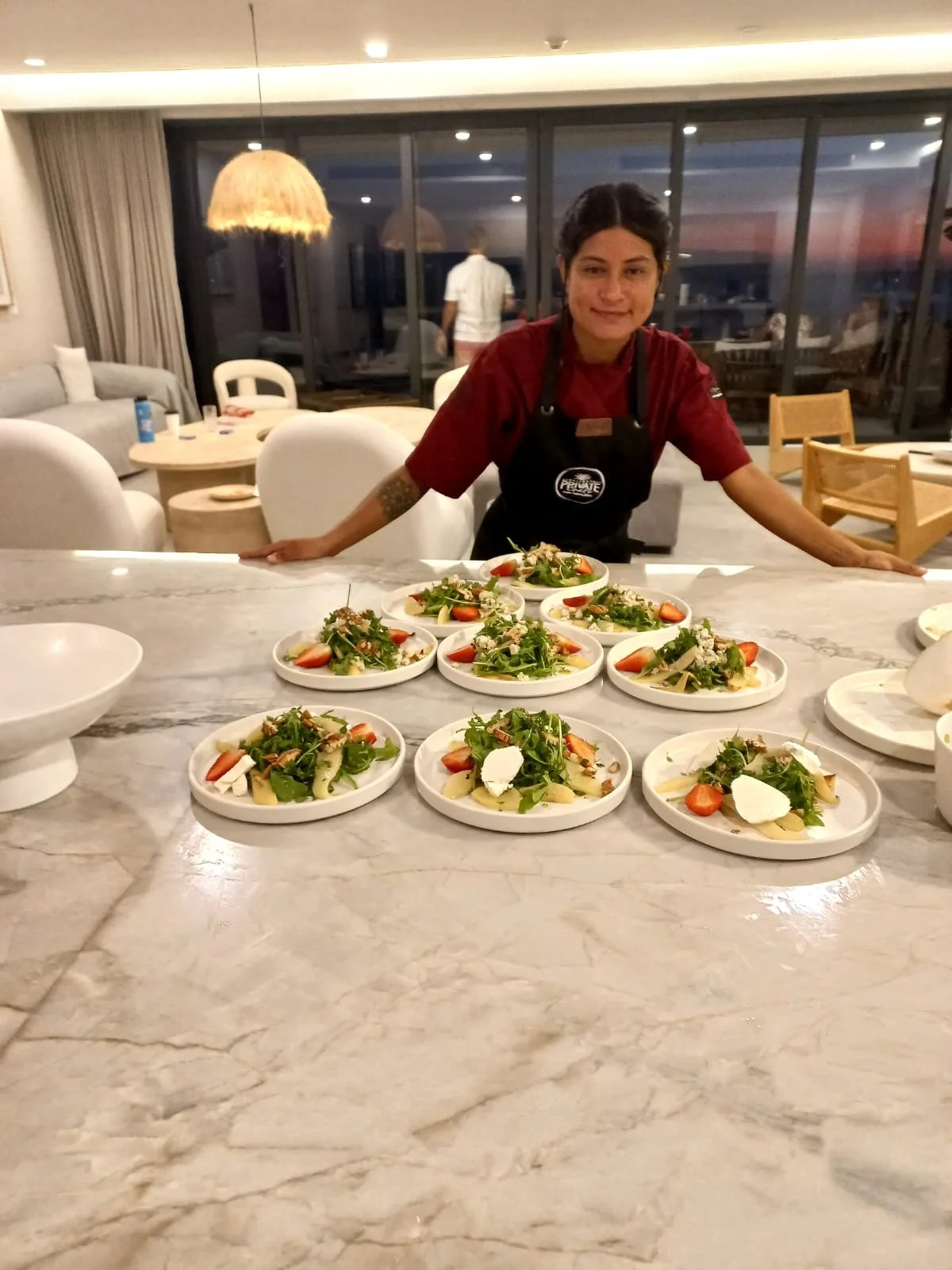 A waitress in a red shirt and black apron stands behind a marble countertop with ten plates of salad topped with strawberries, nuts, cheese, and greens. The restaurant interior features a large window with a sunset view, a seating area with white cha