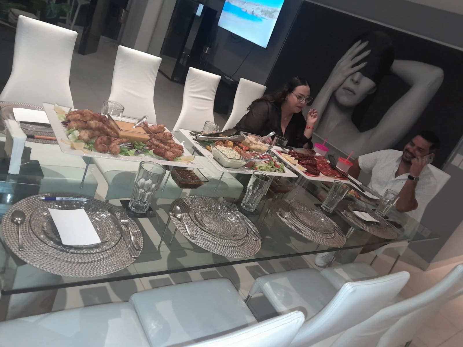A group of people sitting at a dining table with various dishes including fried chicken, salads, and meats, in a modern dining room with white chairs and a large artwork on the wall.