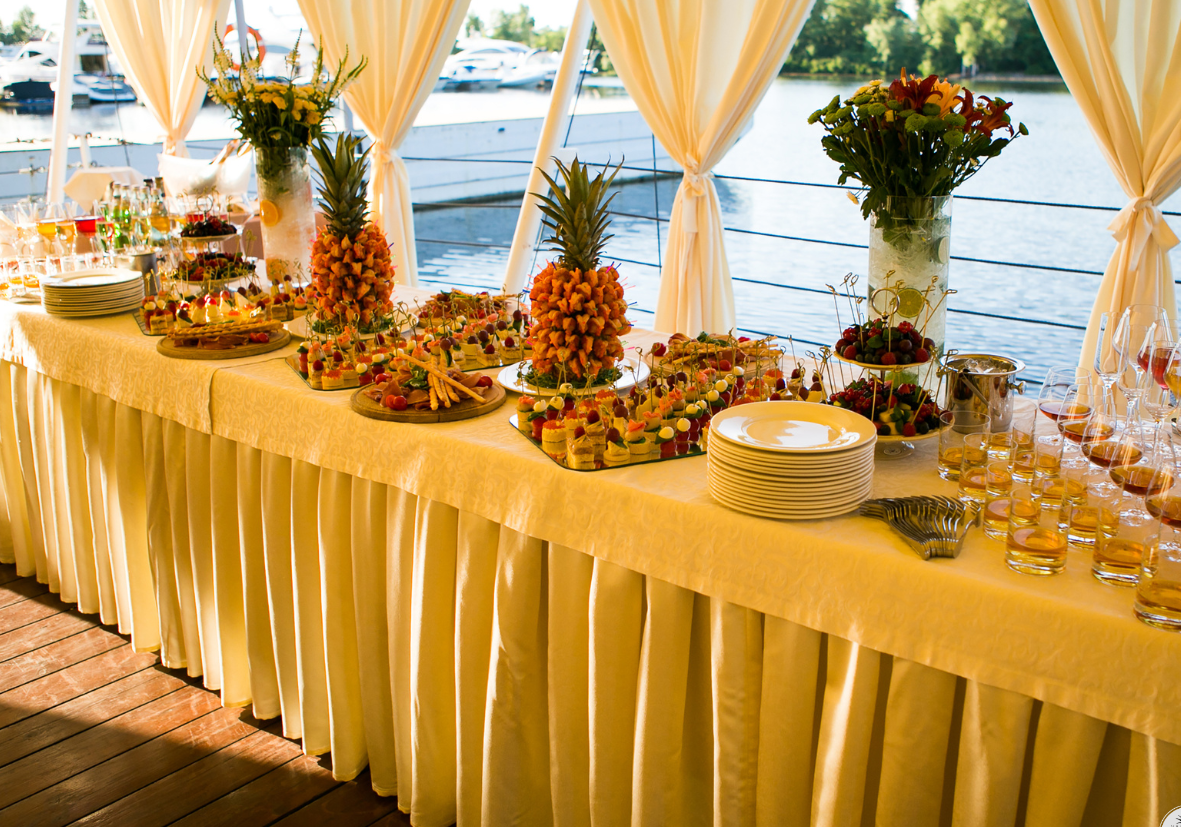 Buffet table with tropical fruit displays, desserts, flower arrangements, and drinks near a lake with boats, inside a tent with cream-colored curtains.