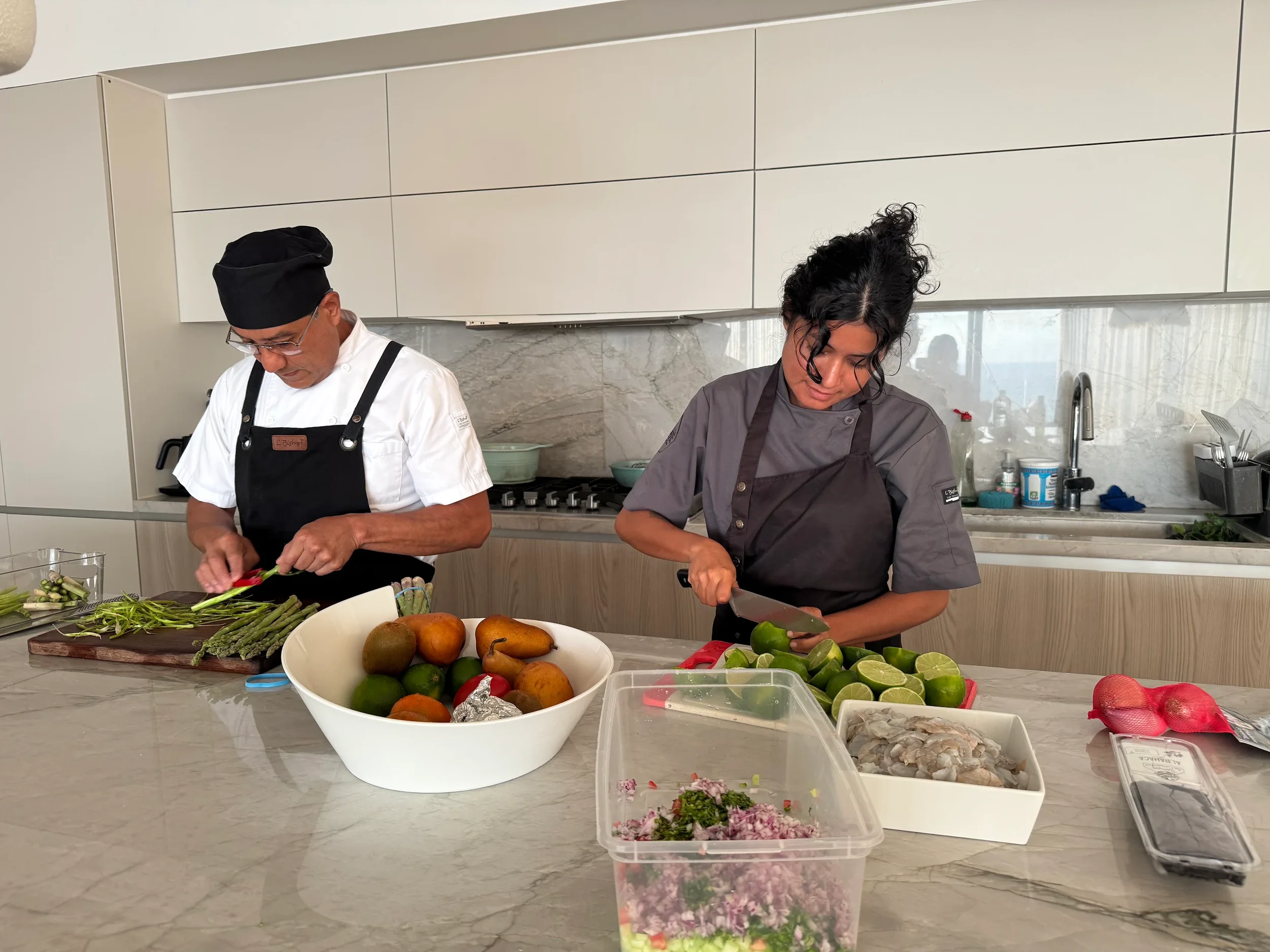 Two people in chef uniforms preparing vegetables in a modern kitchen with white cabinets and a marble backsplash. There is a bowl of pears and limes, chopped vegetables, and various ingredients on the counter.
