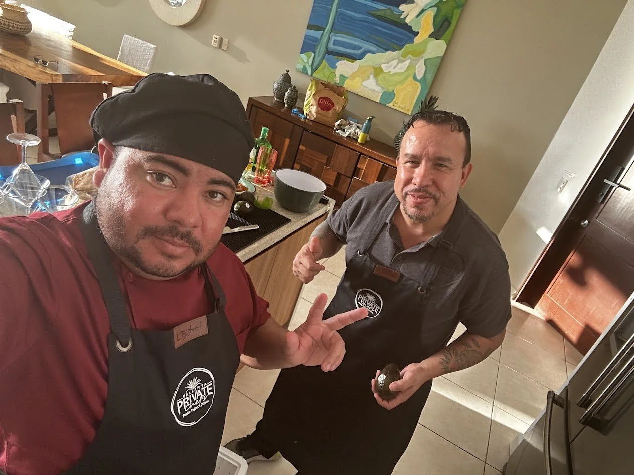 Two men in a kitchen taking a selfie. One is wearing a black chef's hat and red shirt, the other is wearing a gray shirt and apron with a logo. They are smiling, and the man in the apron is holding an avocado.