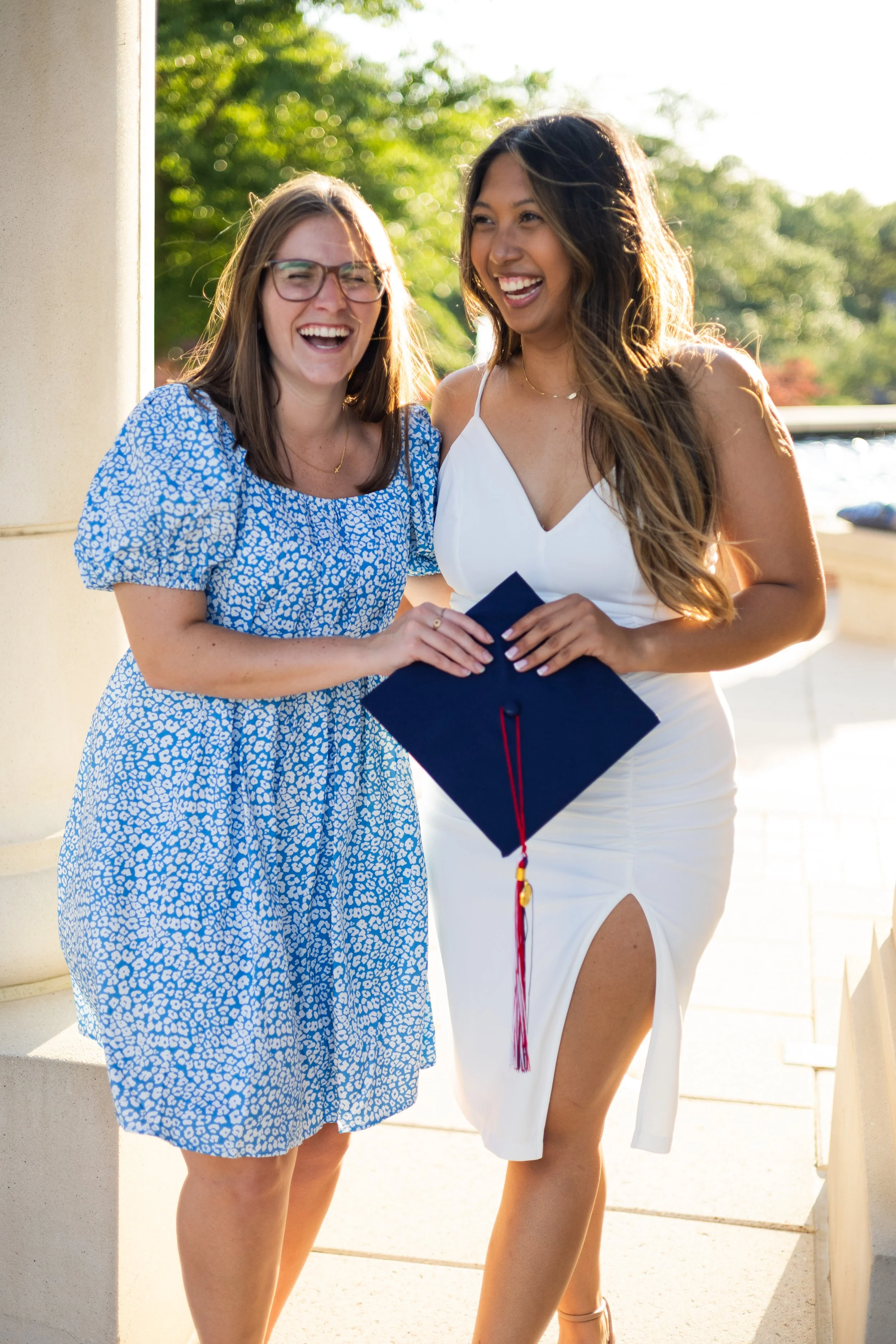 Two women standing outside, smiling and sharing a joyful moment. One is holding a diploma and wearing a white dress, and the other is wearing a blue patterned dress and glasses.