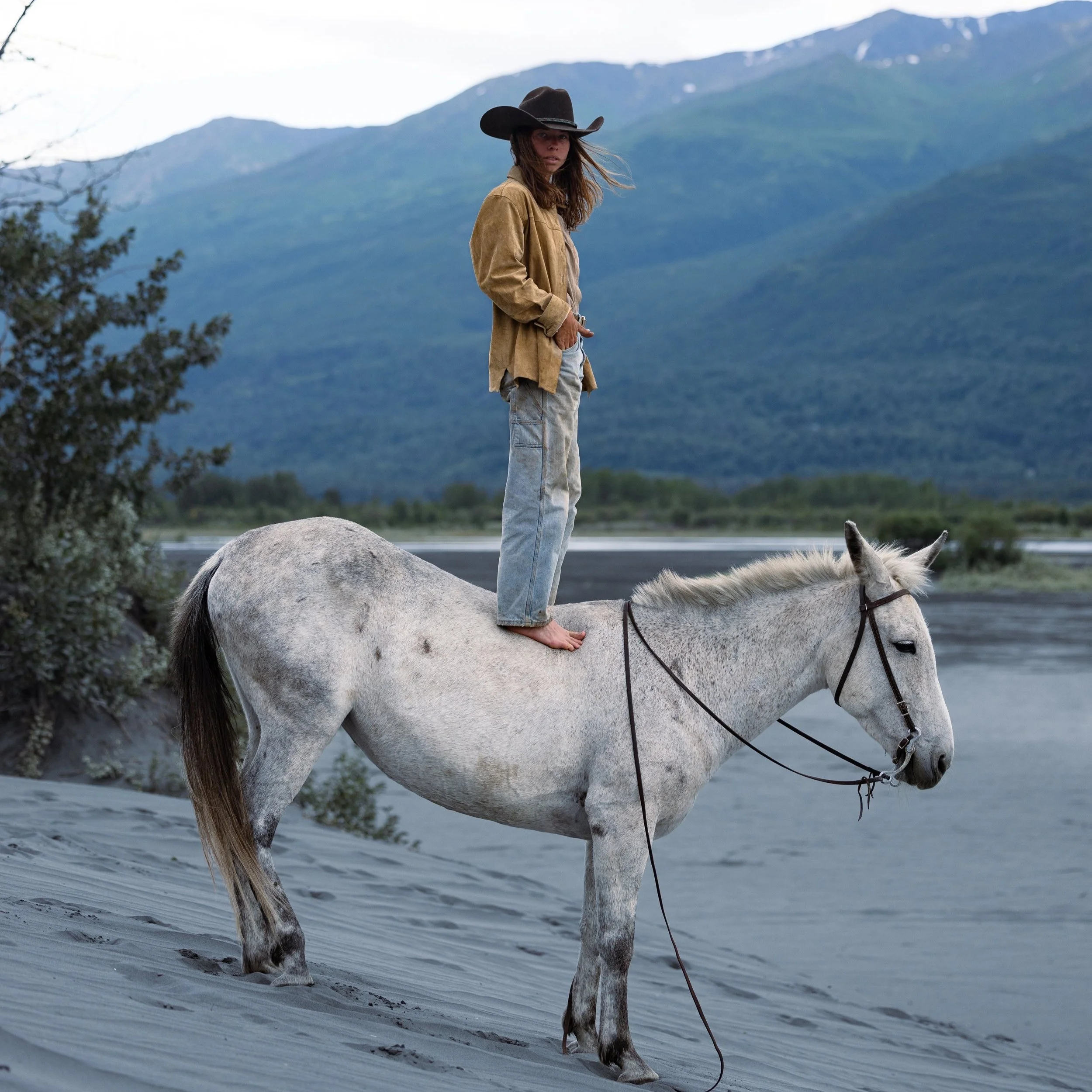 A woman in cowboy hat and casual clothing walking barefoot on a white horse in a natural outdoor setting with mountains and a body of water in the background.