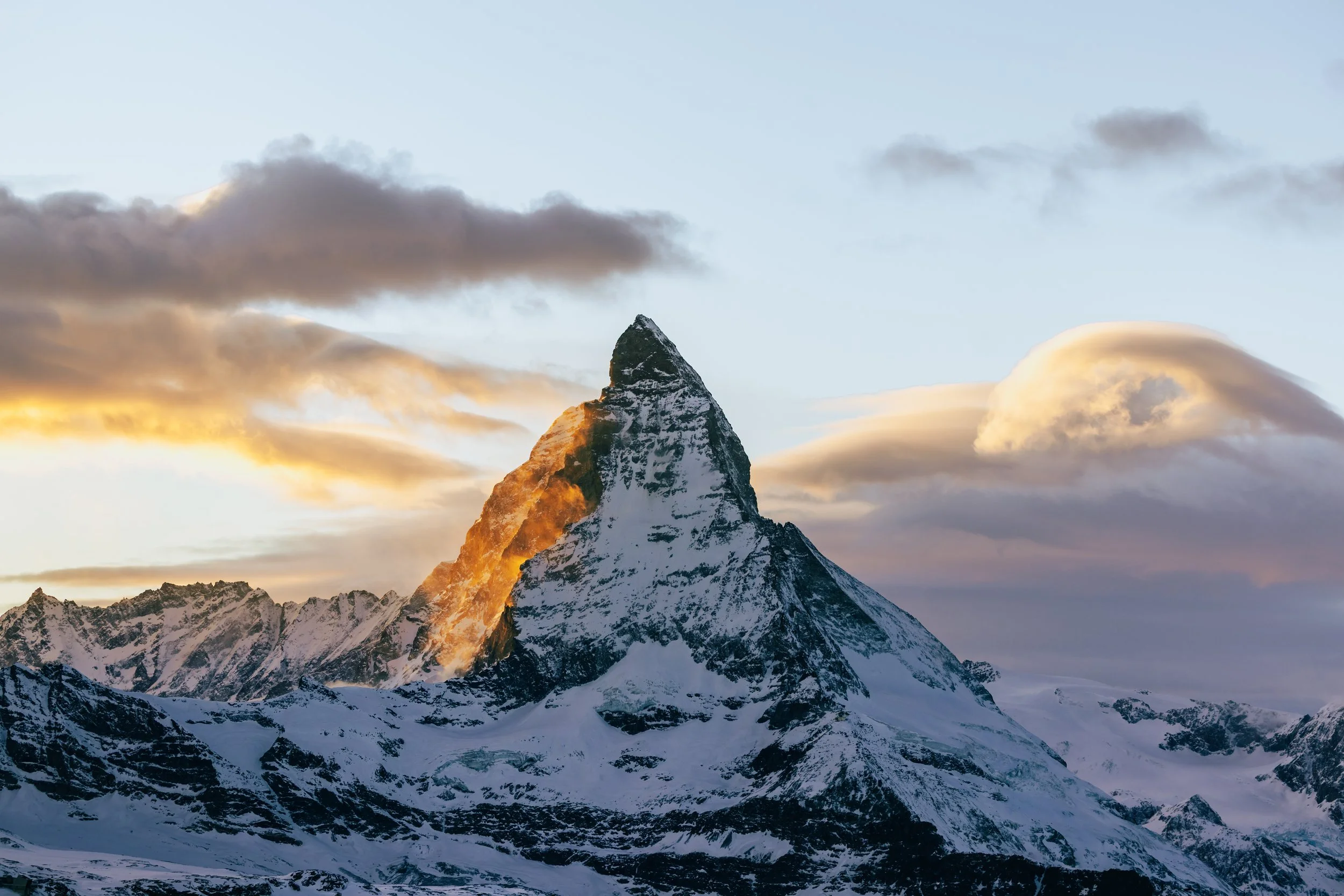 Snow-covered mountain peak with clouds and a partly cloudy sky during sunset or sunrise.