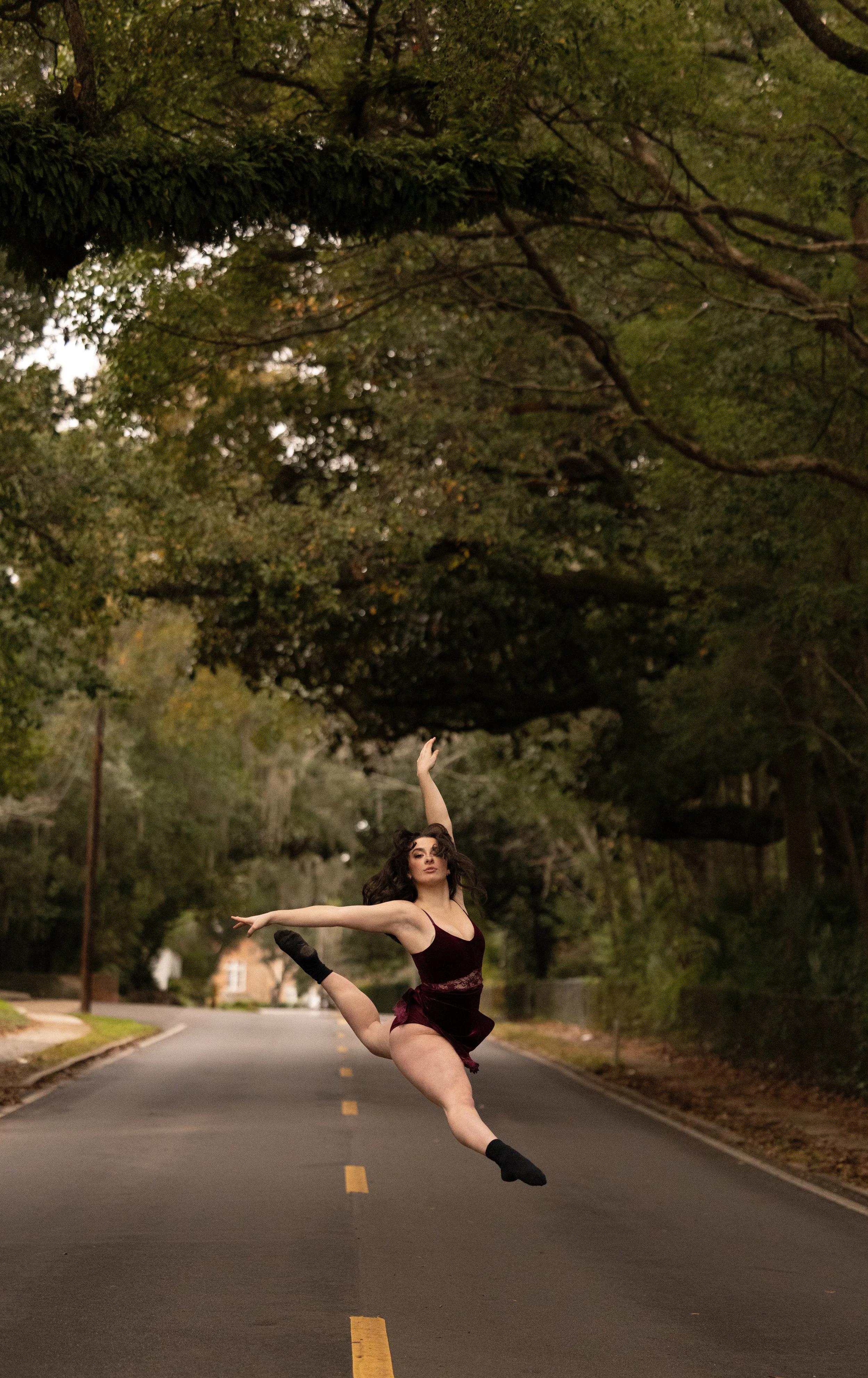 A young woman in a maroon ballet costume performs an elegant ballet pose on an empty residential street lined with trees.