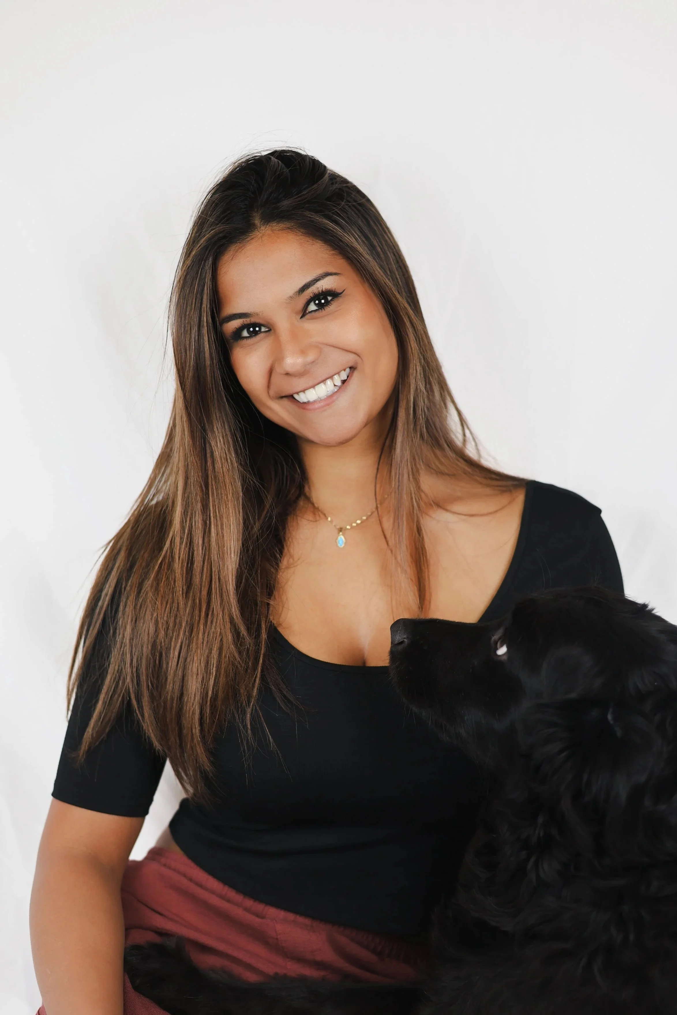 A woman with long brown hair smiling, wearing a black top, and a gold necklace with a blue pendant. She is holding a black dog.