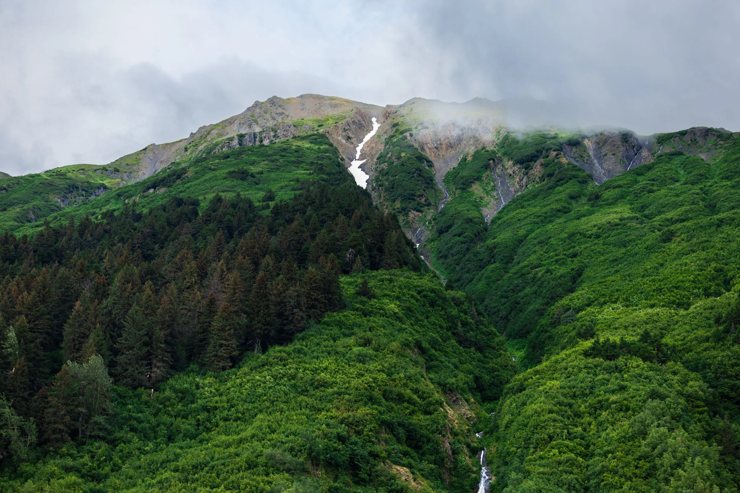 Lush green mountains with patches of snow and a cloudy sky.