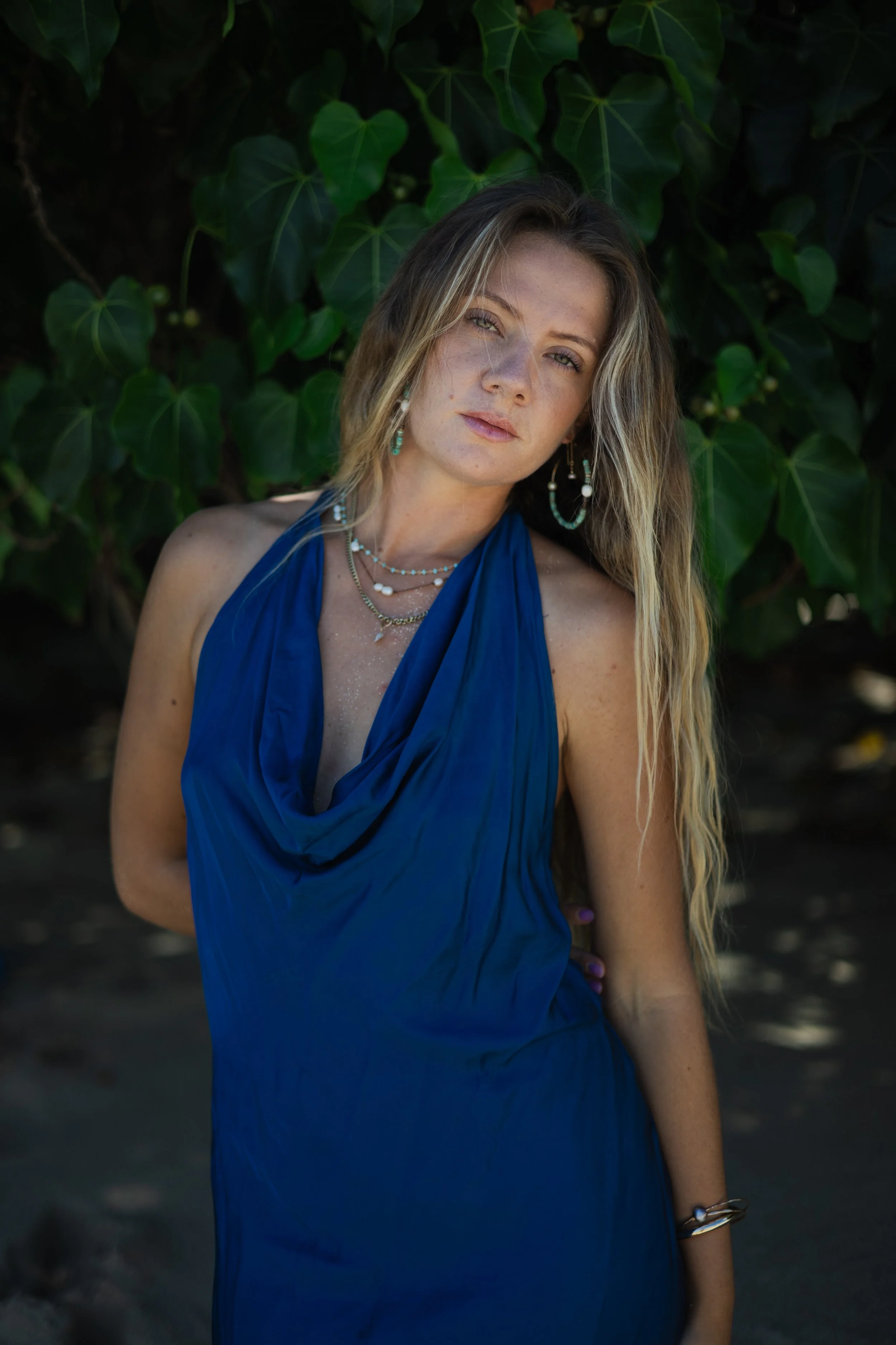 A woman with long, blonde hair wearing a blue dress and jewelry, standing outdoors in front of green foliage.