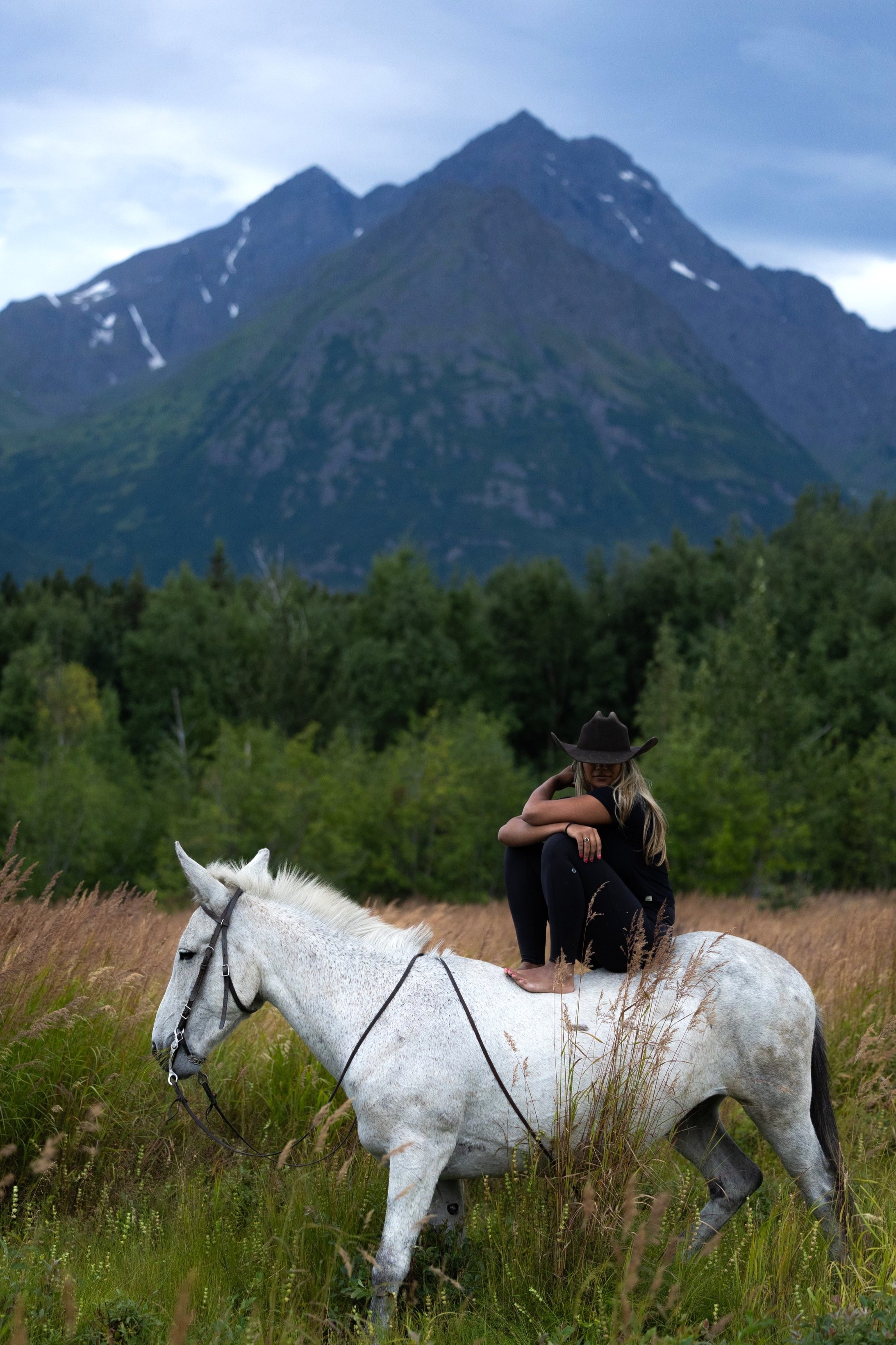 A woman sitting barefoot on a white horse in a grassy field with trees and mountains in the background.
