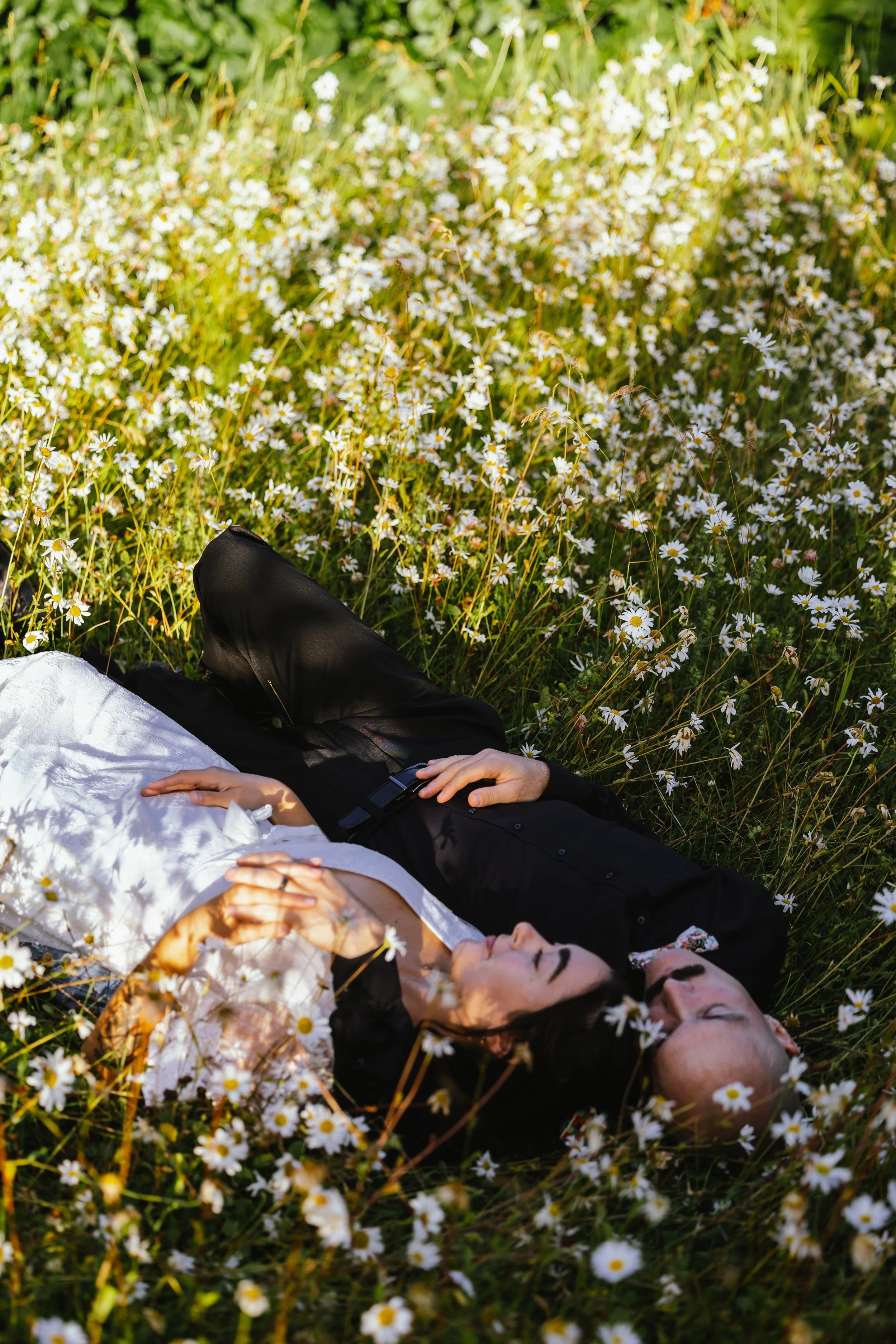 Person lying in a field of white daisies, wearing a white dress and black jacket, with hands resting on stomach and eyes closed.