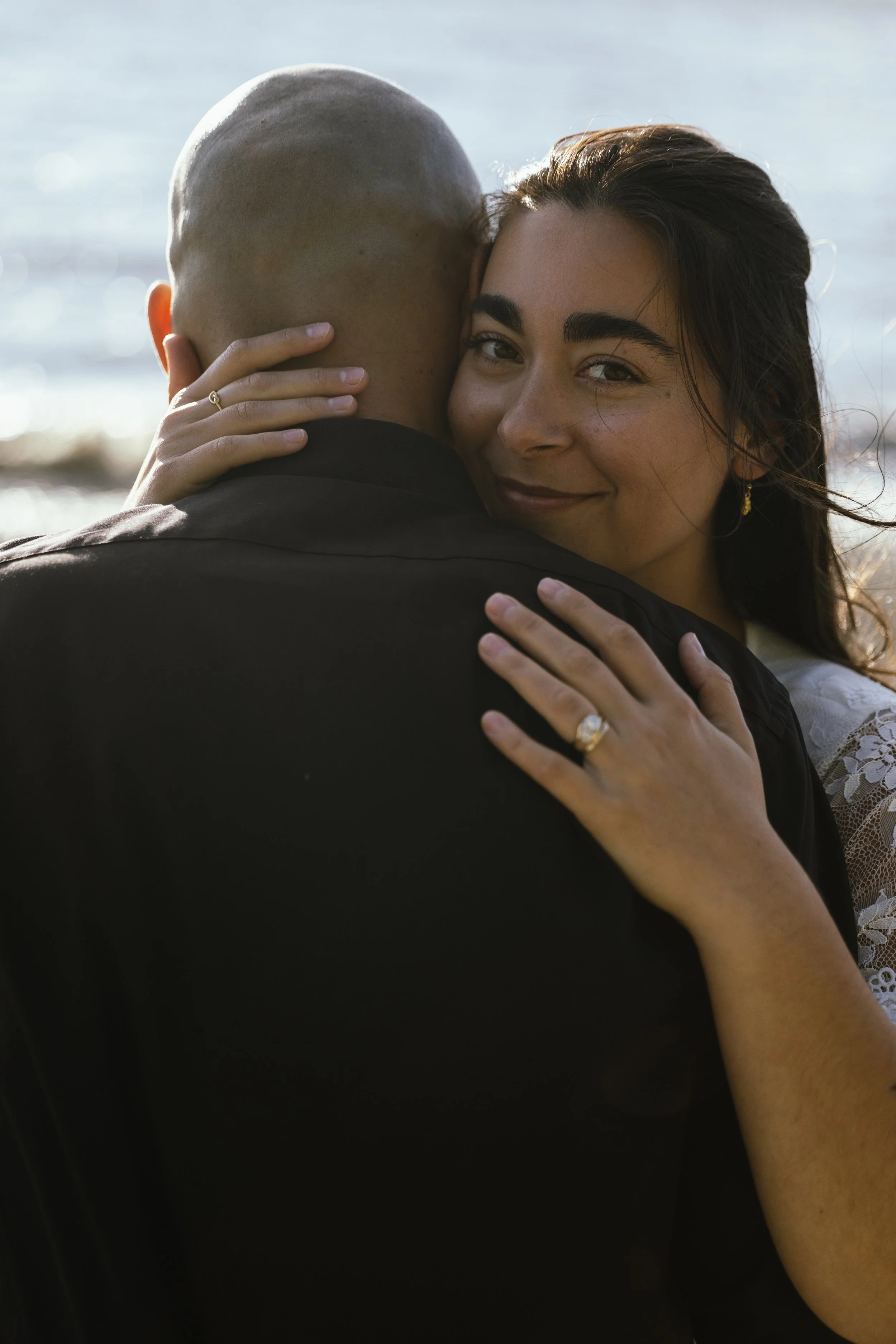 A woman with dark hair and prominent eyebrows smiling while hugging a man whose face is not visible, near a body of water.
