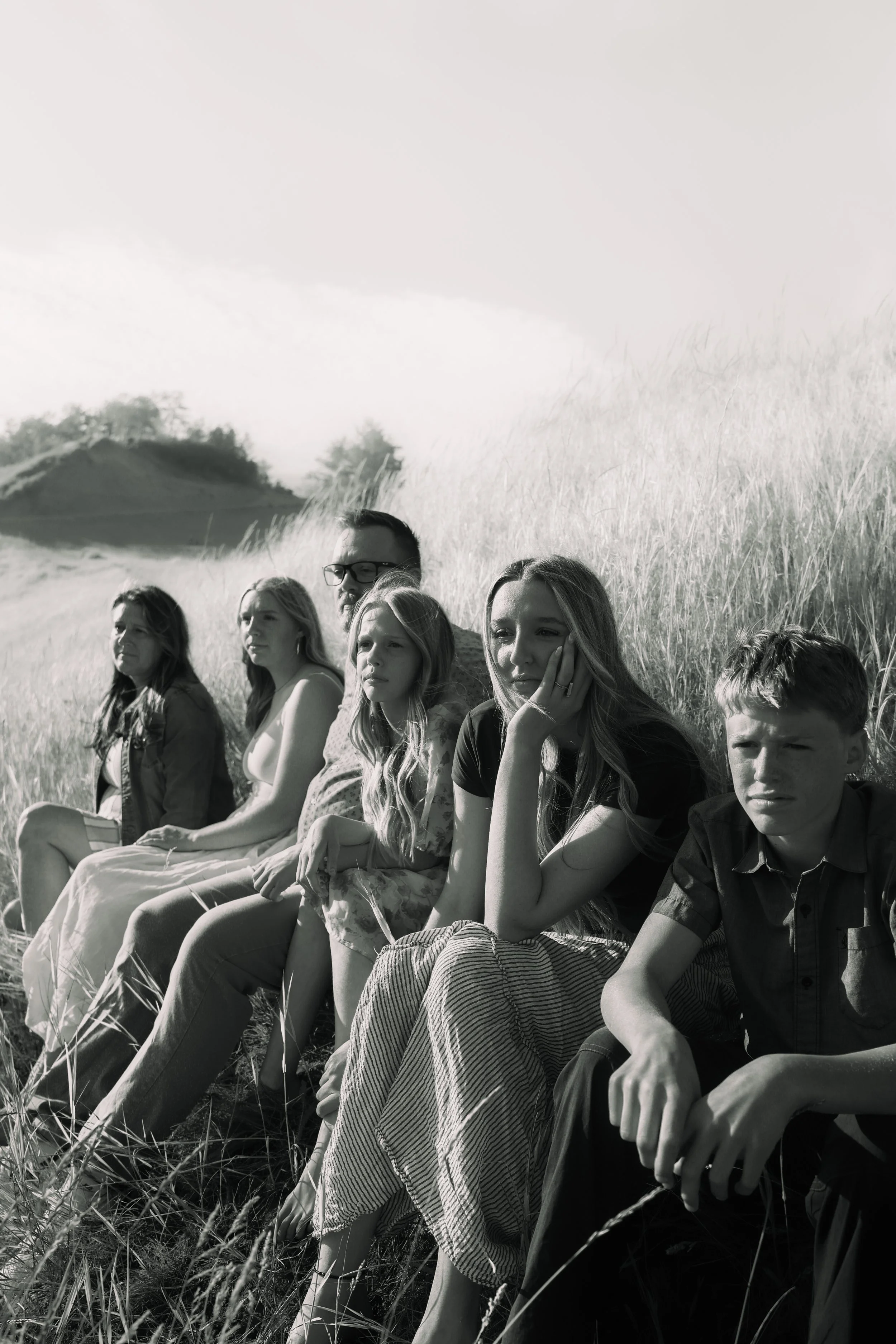 A group of young people sitting on the grass in a field, some with thoughtful expressions, with trees and a hill in the background, black and white photo