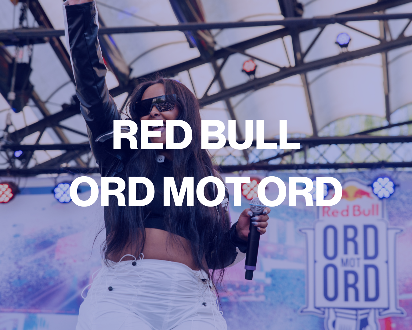 A woman with long dark hair wearing sunglasses and a black leather jacket holding a microphone on an outdoor stage during a Red Bull event. There is a large display with the Red Bull logo and the words "RED BUL" and "ORD MOT ORD" in the background, with stage lighting above.