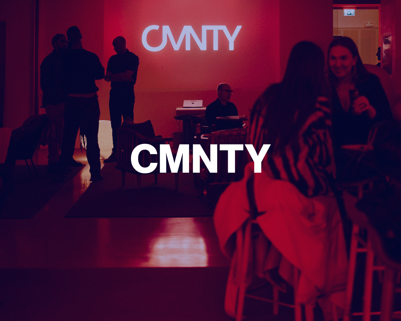 People socializing at an indoor event with a large illuminated sign displaying 'CMNTY' in the background