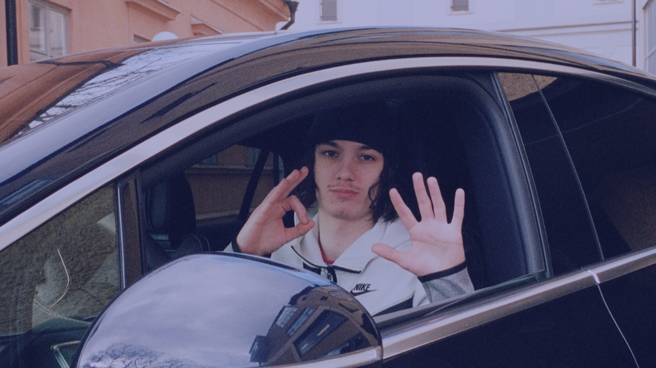 Young man with curly dark hair and light skin sitting inside a black car, making an OK hand gesture with one hand and waving with the other, looking at the camera through the window.