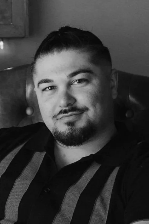 A young man with short dark hair and a goatee, sitting on a leather bench, wearing a black and gray striped polo shirt.