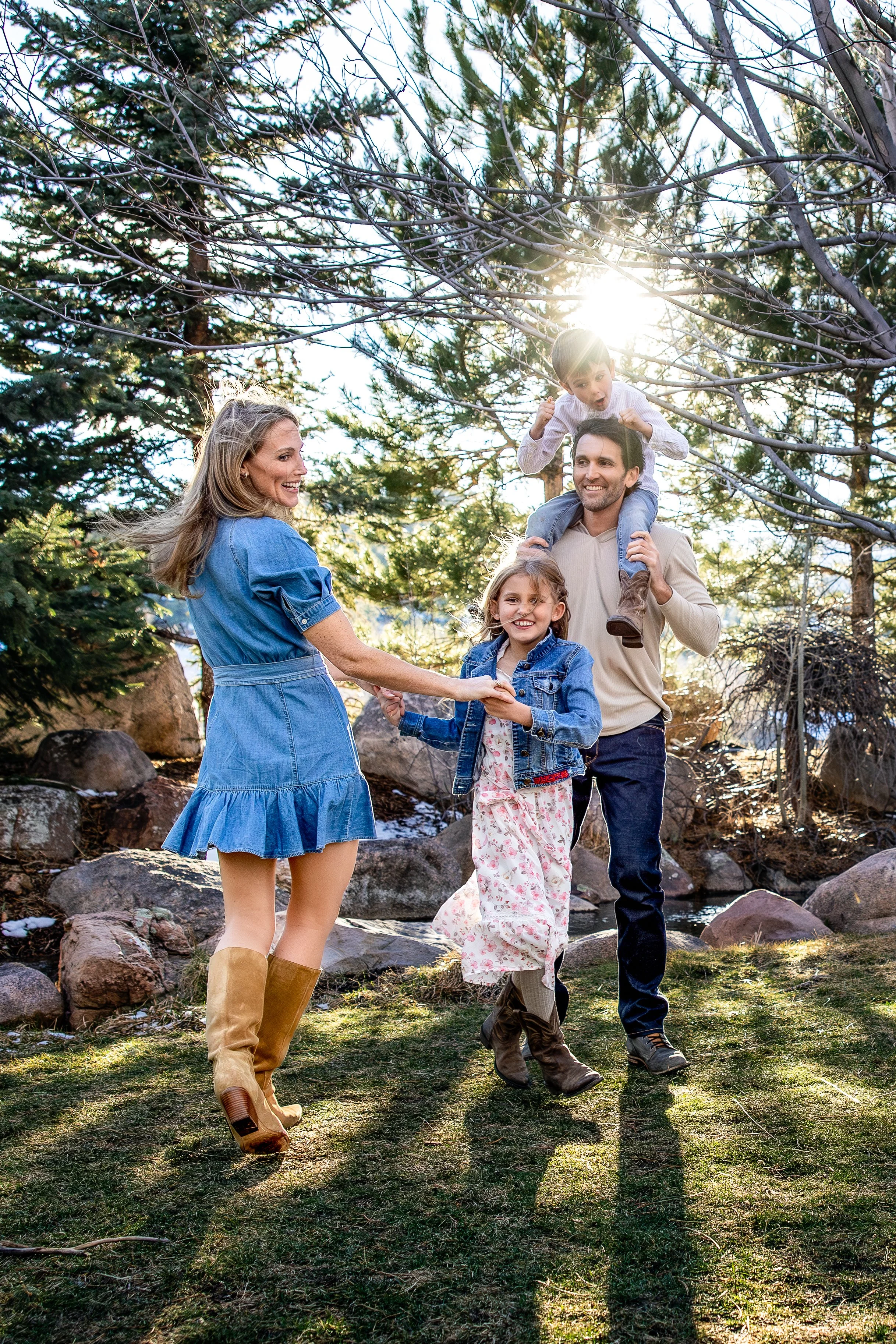 A family of four outdoors in a wooded area, with the sun shining through trees. The father carries a young boy on his shoulders, and the mother holds hands with a young girl. All are smiling and enjoying the moment.