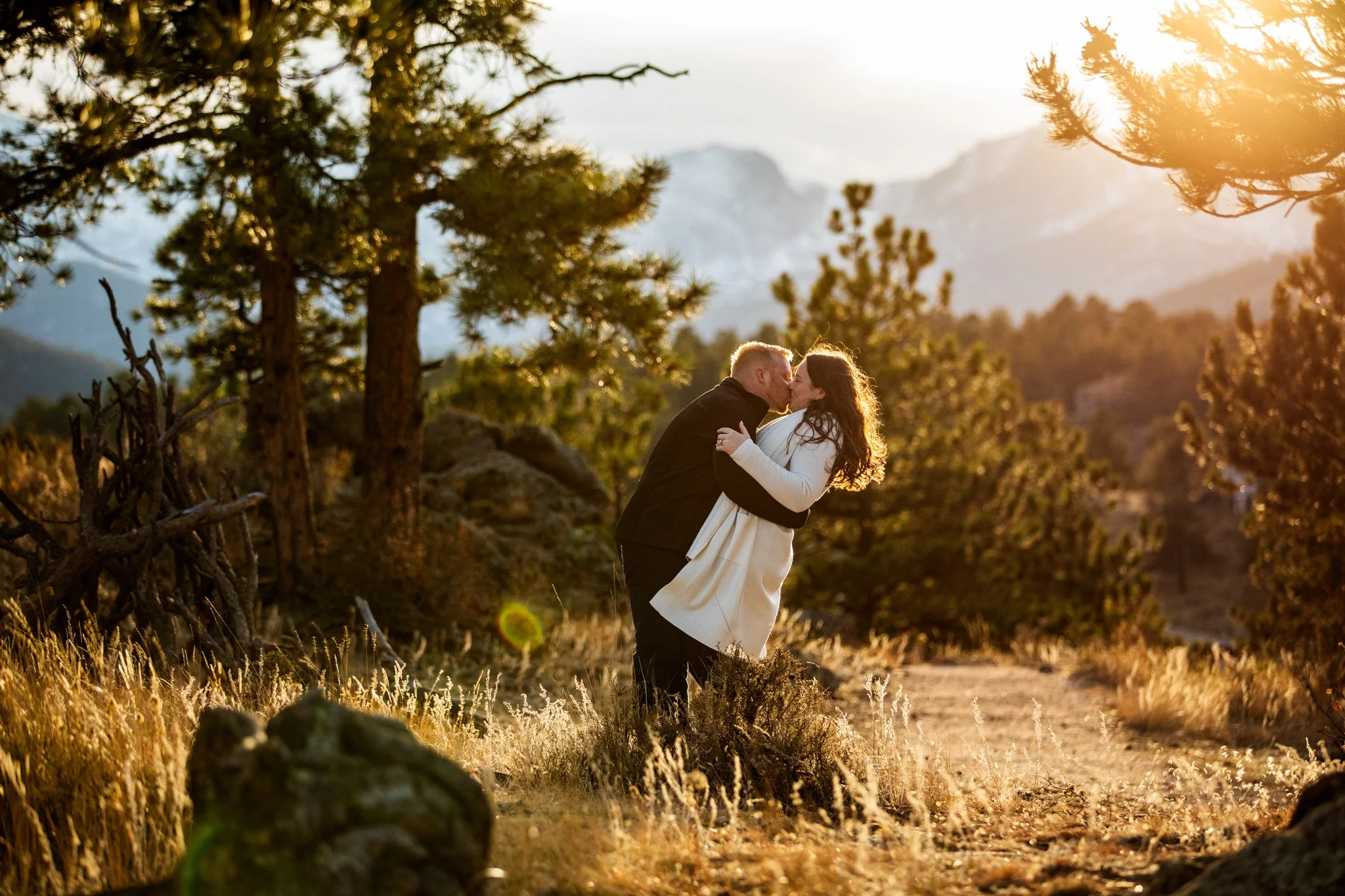 A couple kissing outdoors on a sunny day in a mountainous, forested area with trees and rocks, during sunset.