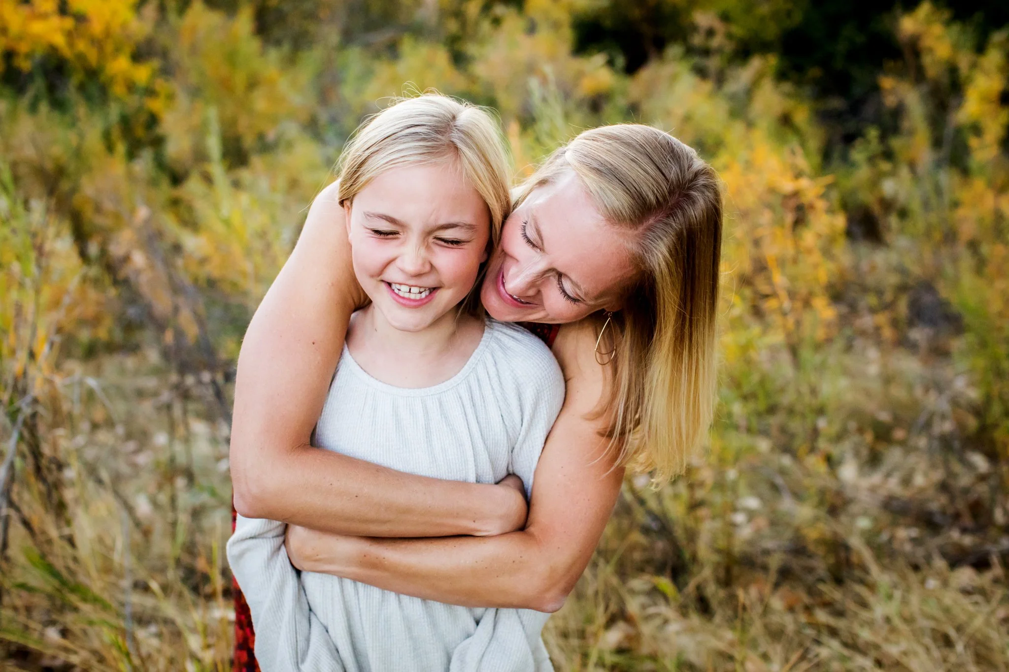A woman hugging a smiling girl outdoors with fall foliage in the background.