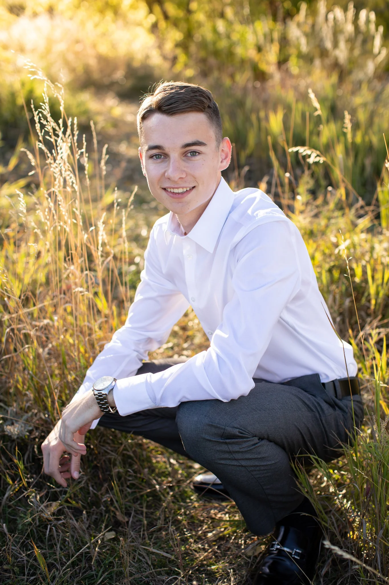 High school portrait photographer takes a pictures of a young man in a slacks and a white collared shirt kneeling down in a field.