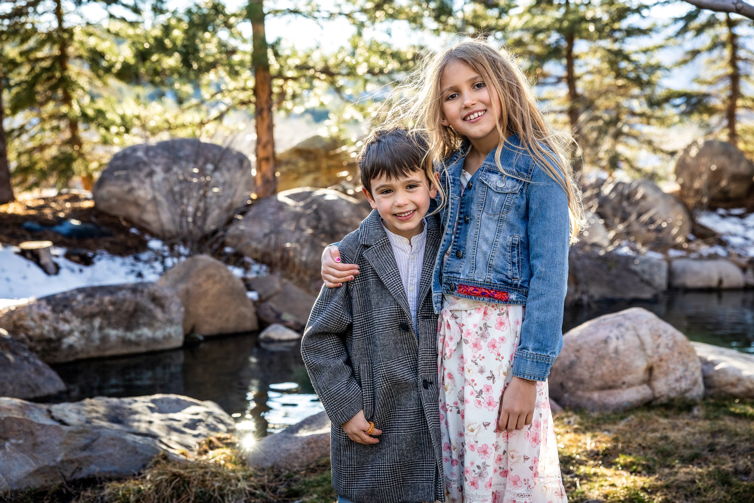 Two children, a boy and a girl, standing outdoors near a stream with rocks and pine trees in the background, smiling and embracing.