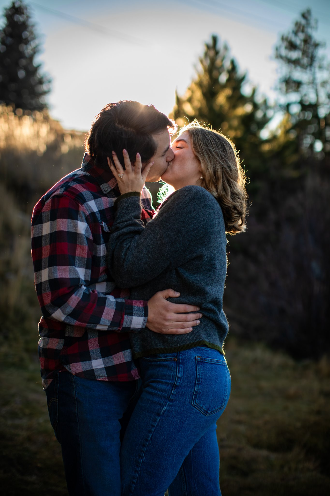 A couple kissing outdoors during sunset, dressed casually, with trees and a natural landscape in the background.
