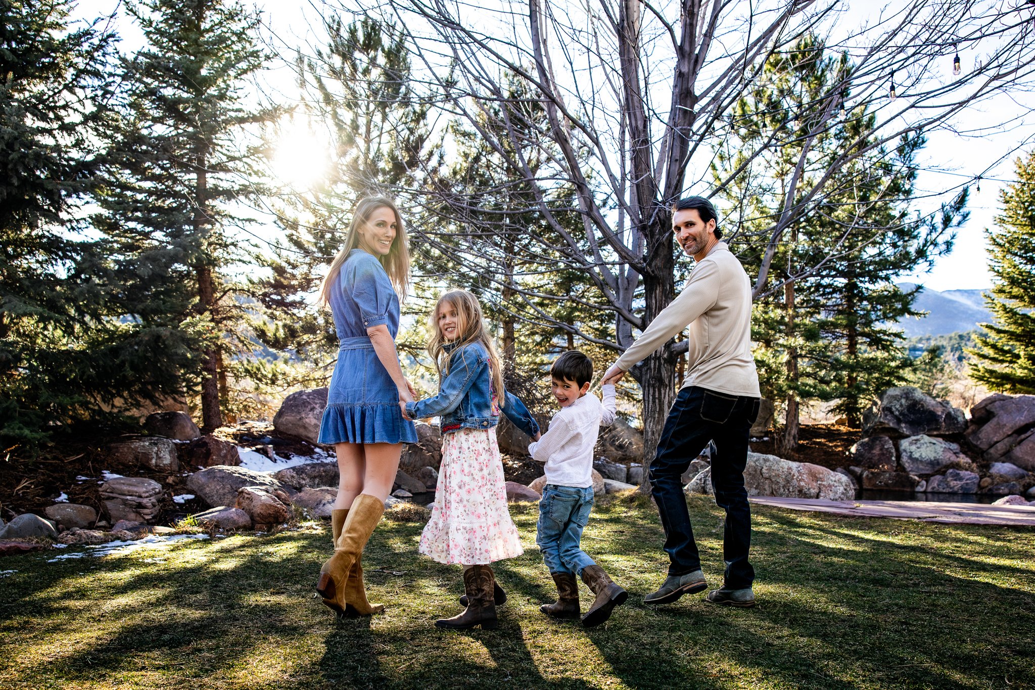 A family of five, including two adults and three children, holding hands and playing outdoors in a forested area during the daytime. Sunlight filters through trees in the background.