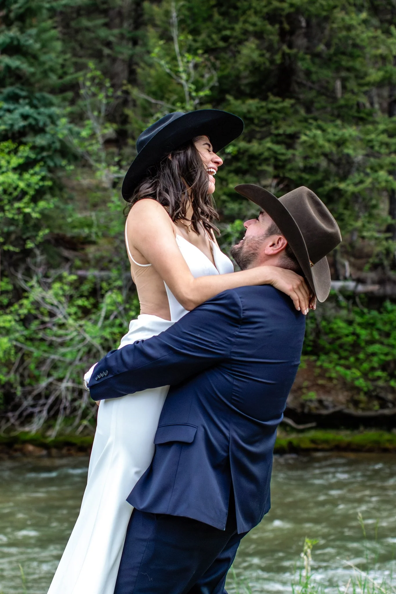 A man lifting a woman in a white dress, both wearing cowboy hats, outdoors near a river with green trees in the background, smiling and looking at each other.