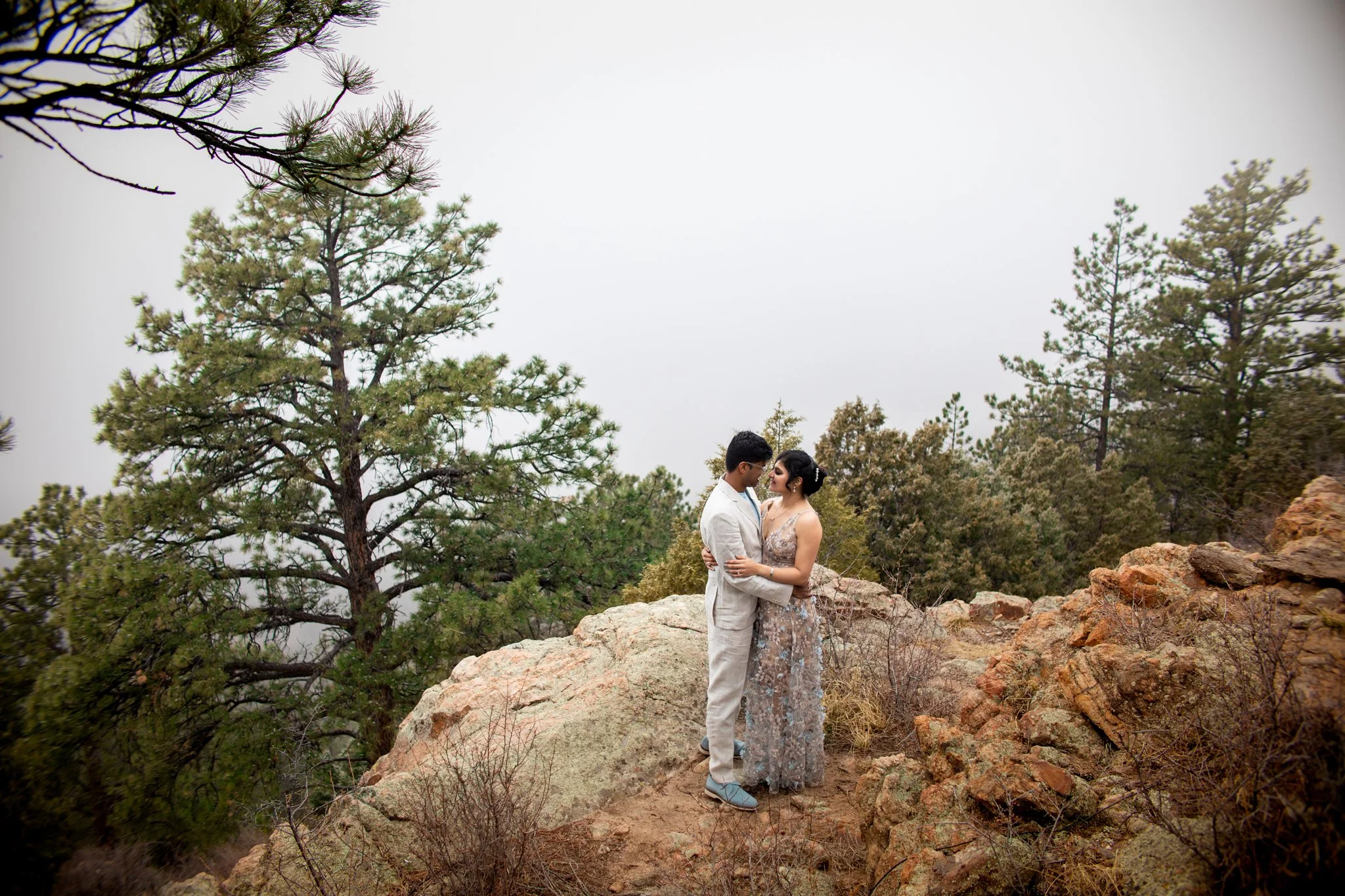 A couple dressed in wedding attire embracing on rocky terrain surrounded by pine trees in a mountainous area under an overcast sky.